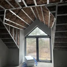 Poddasze w trakcie ocieplania: widoczne wełna mineralna między metalowymi profilami konstrukcji sufitu i ścian, okno trójkątne, styropian na podłodze, surowy beton.