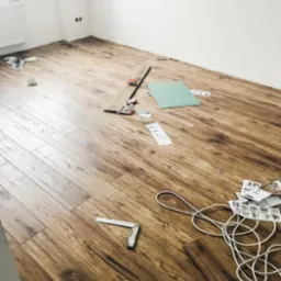 Proces montażu paneli podłogowych w pomieszczeniu z białymi ścianami, widoczne narzędzia: kątownik, miarka, fragment arkusza zielonej płyty, oraz resztki opakowań po panelach.