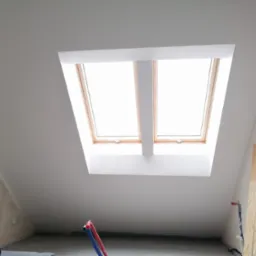 Poddasze po remoncie z oknem dachowym; świeżo pomalowane ściany i sufit w kolorze białym, widoczne rury instalacyjne i płyty budowlane przy ścianie.