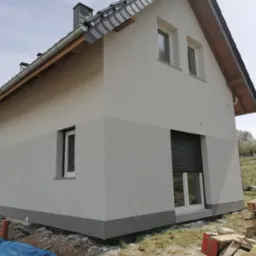 Nowoczesny dom jednorodzinny z jasną elewacją w dwóch odcieniach szarości, dach pokryty ciemną dachówką, okno z roletą zewnętrzną, widoczne materiały budowlane wokół domu.