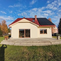 SEBUD - Jasny dom jednorodzinny z czerwonym dachem, czarnymi panelami fotowoltaicznymi i dużymi oknami tarasowymi. Przed nim betonowy taras, zielony trawnik i jesienne drzewa pod błękitnym niebem.