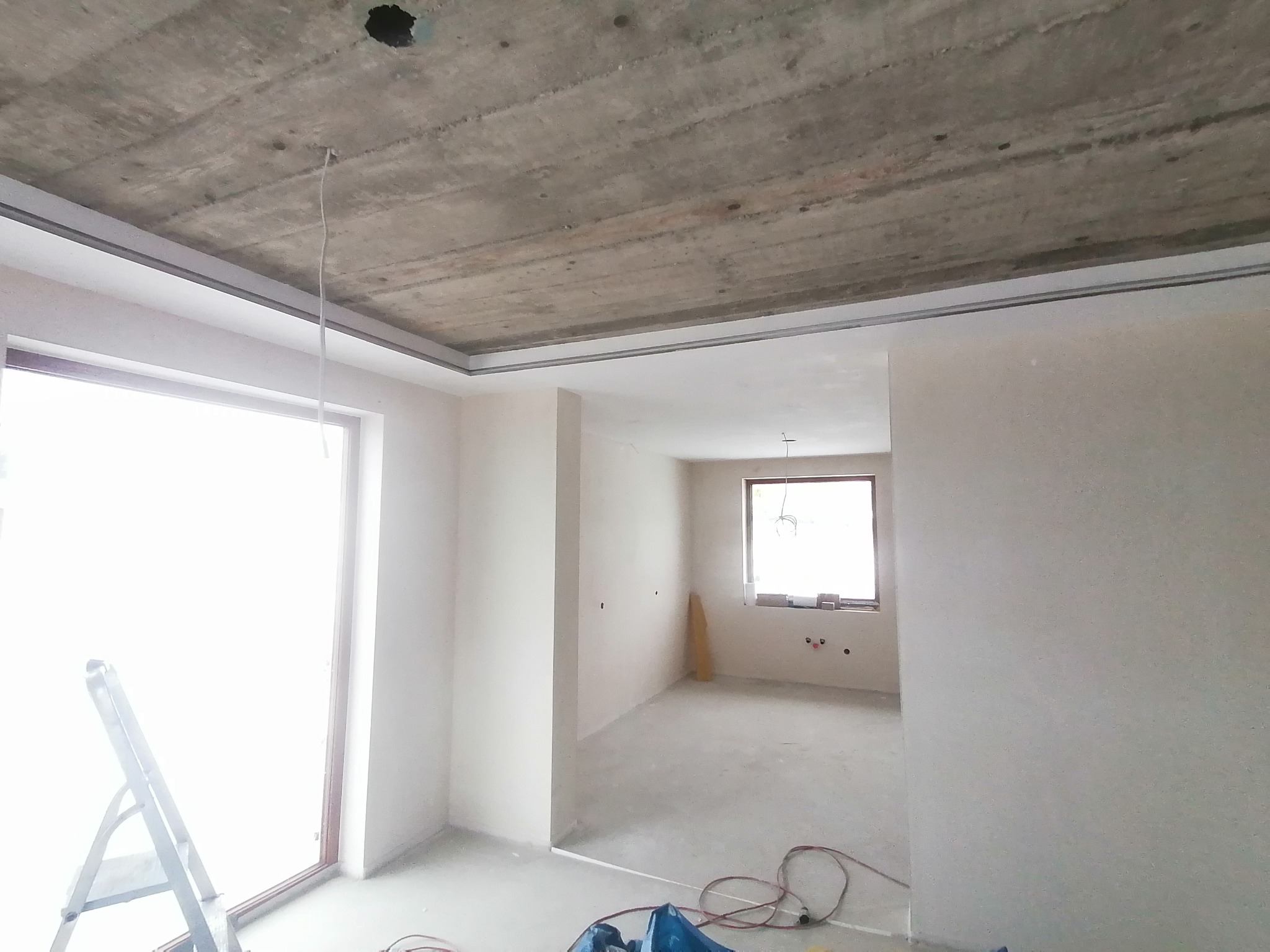 Surowy betonowy sufit z widocznymi śladami szalunków, białe ściany w trakcie wykańczania, aluminiowe profile sufitowe, drabina oparta o ścianę, widok na sąsiednie pomieszczenie z oknem.