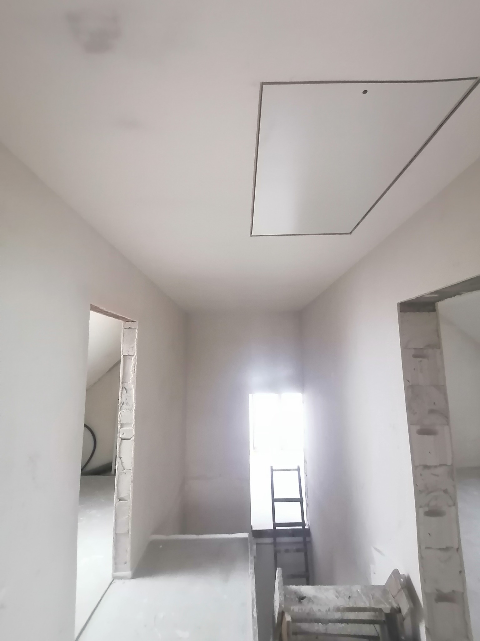 Korytarz w trakcie prac wykończeniowych, widoczne surowe ściany, schody i drabina prowadząca do okna dachowego z klapą rewizyjną w suficie.