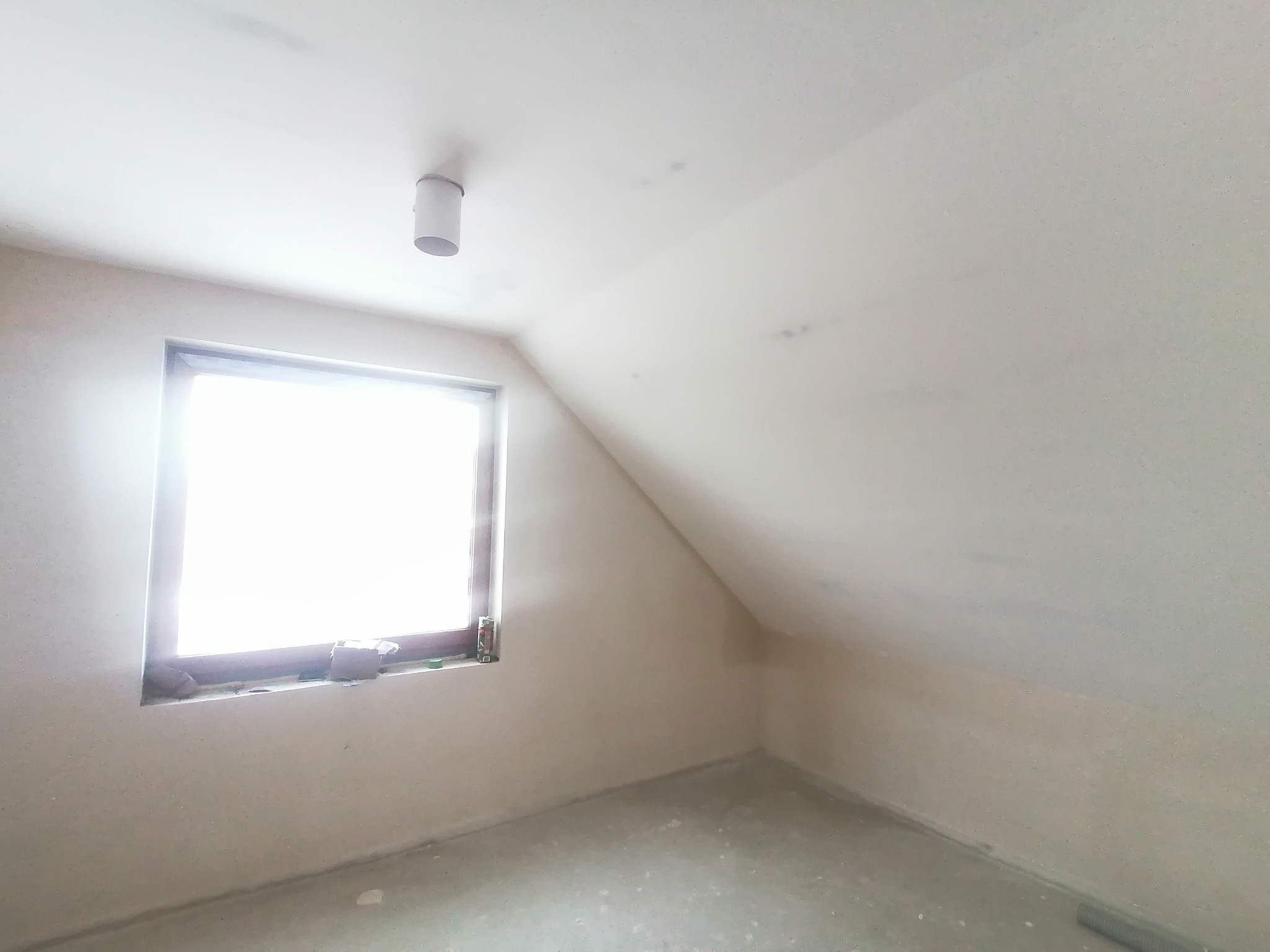 Poddasze po nałożeniu gładzi, widoczne okno z drobnymi przedmiotami na parapecie, sufit ze skośną ścianą i lampą.