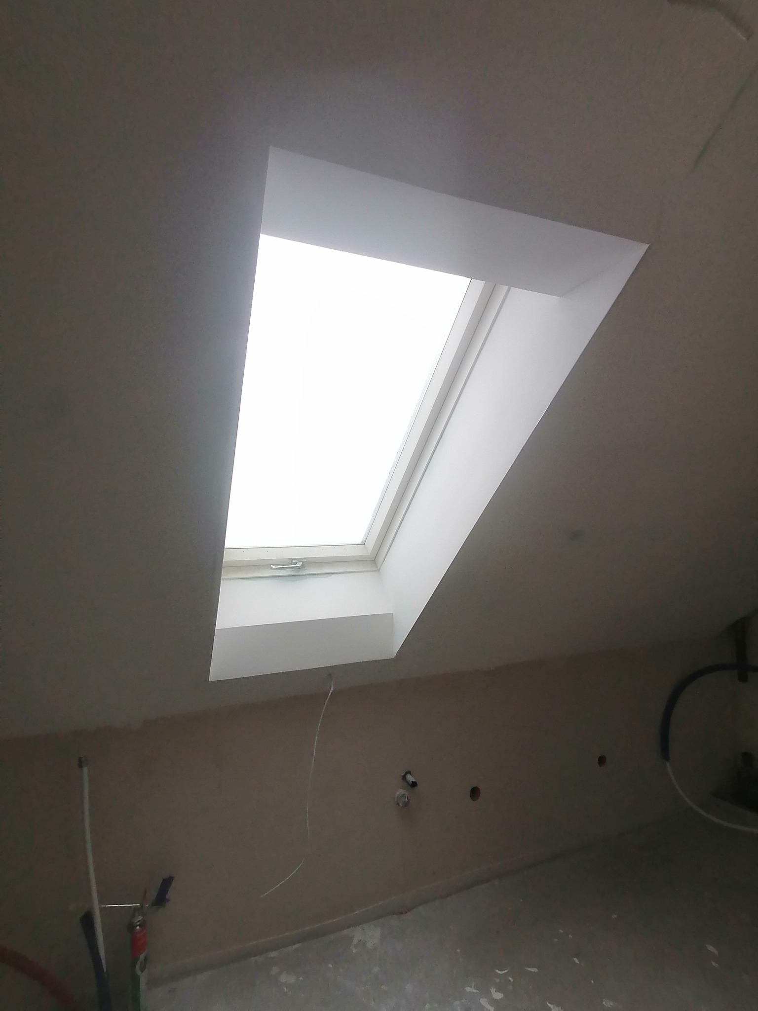 Montaż okna dachowego w skośnym suficie, widoczne niedokończone gładzie na ścianach i podłodze, instalacja elektryczna w tle.