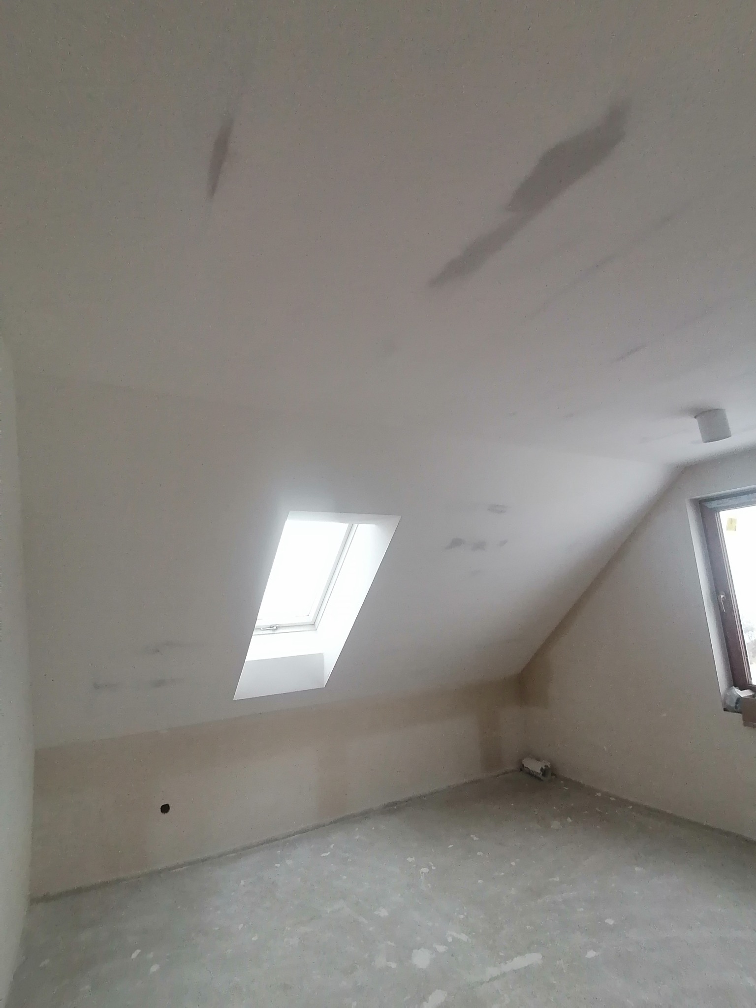 Poddasze po nałożeniu gładzi, widoczne zacieki na suficie i ścianach, okno dachowe oraz okno pionowe po prawej stronie.