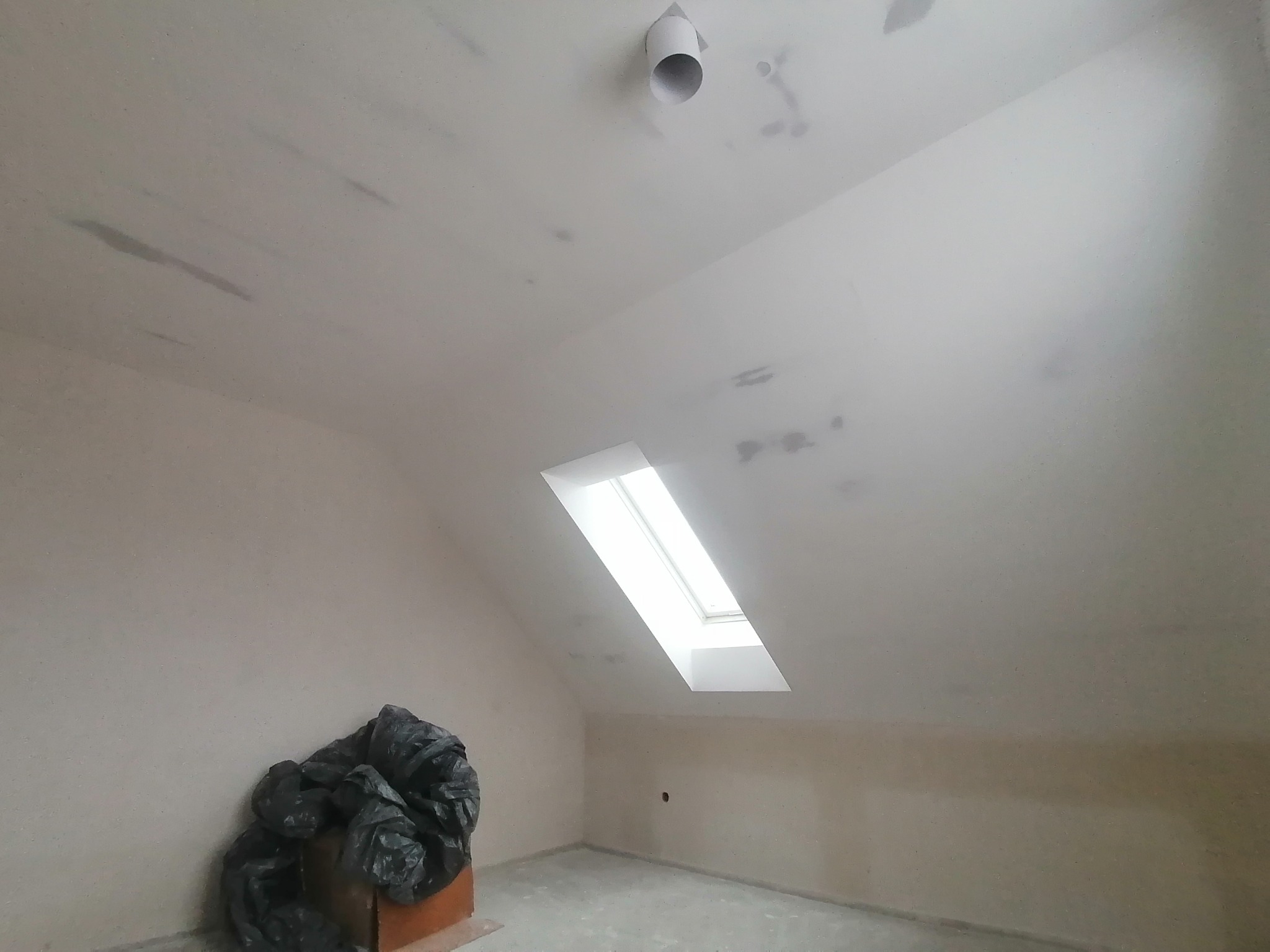 Poddasze po nałożeniu wstępnej warstwy gładzi, widoczne ślady szpachlowania na suficie, okno dachowe, wystający fragment rury wentylacyjnej i worki z odpadami budowlanymi w rogu pomieszczenia.