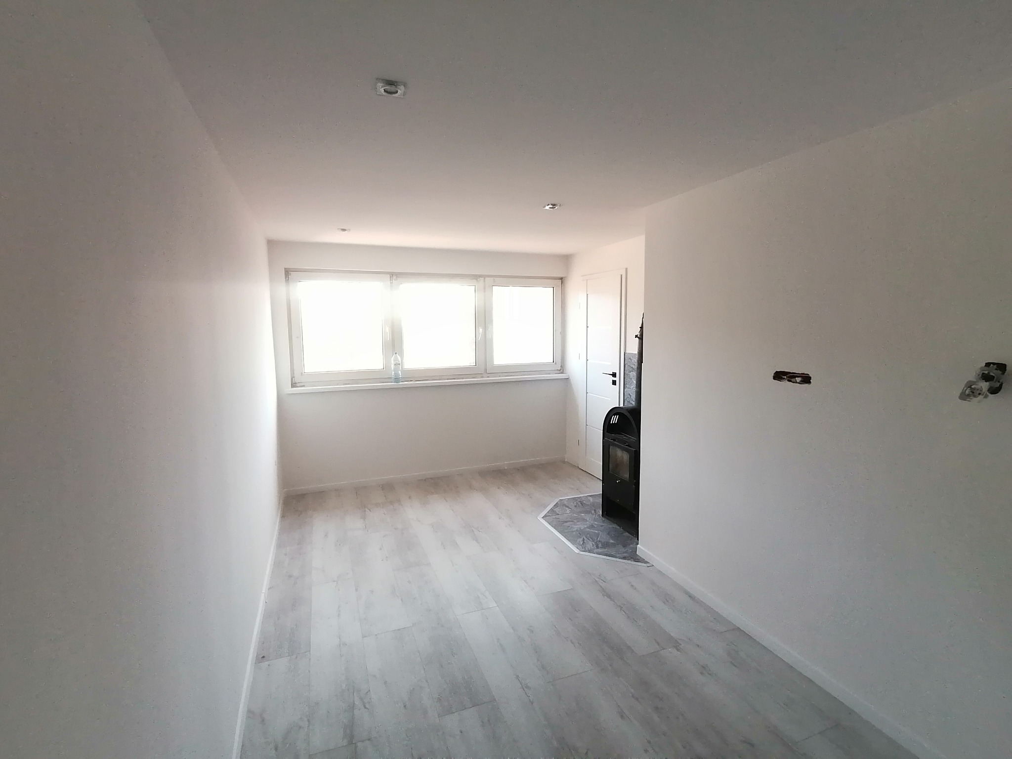 Wnętrze odnowionego pokoju z jasnymi ścianami, panelami podłogowymi imitującymi drewno i czarnym piecem wolnostojącym na tle białych drzwi.