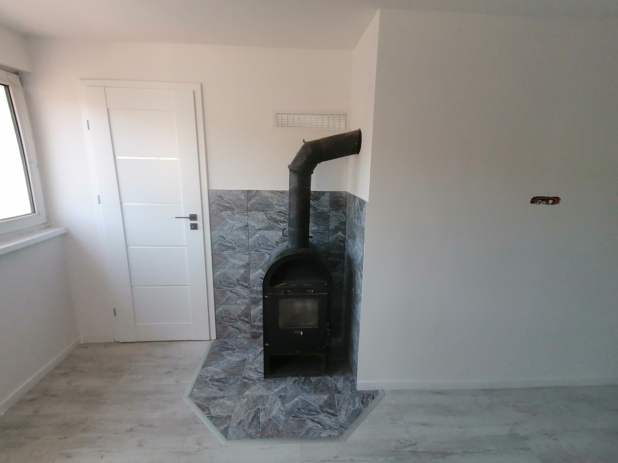 Wnętrze pokoju z zamontowanym czarnym piecem kominkowym, obudowanym płytkami imitującymi kamień, białe ściany, białe drzwi z przeszkleniami, jasna podłoga.