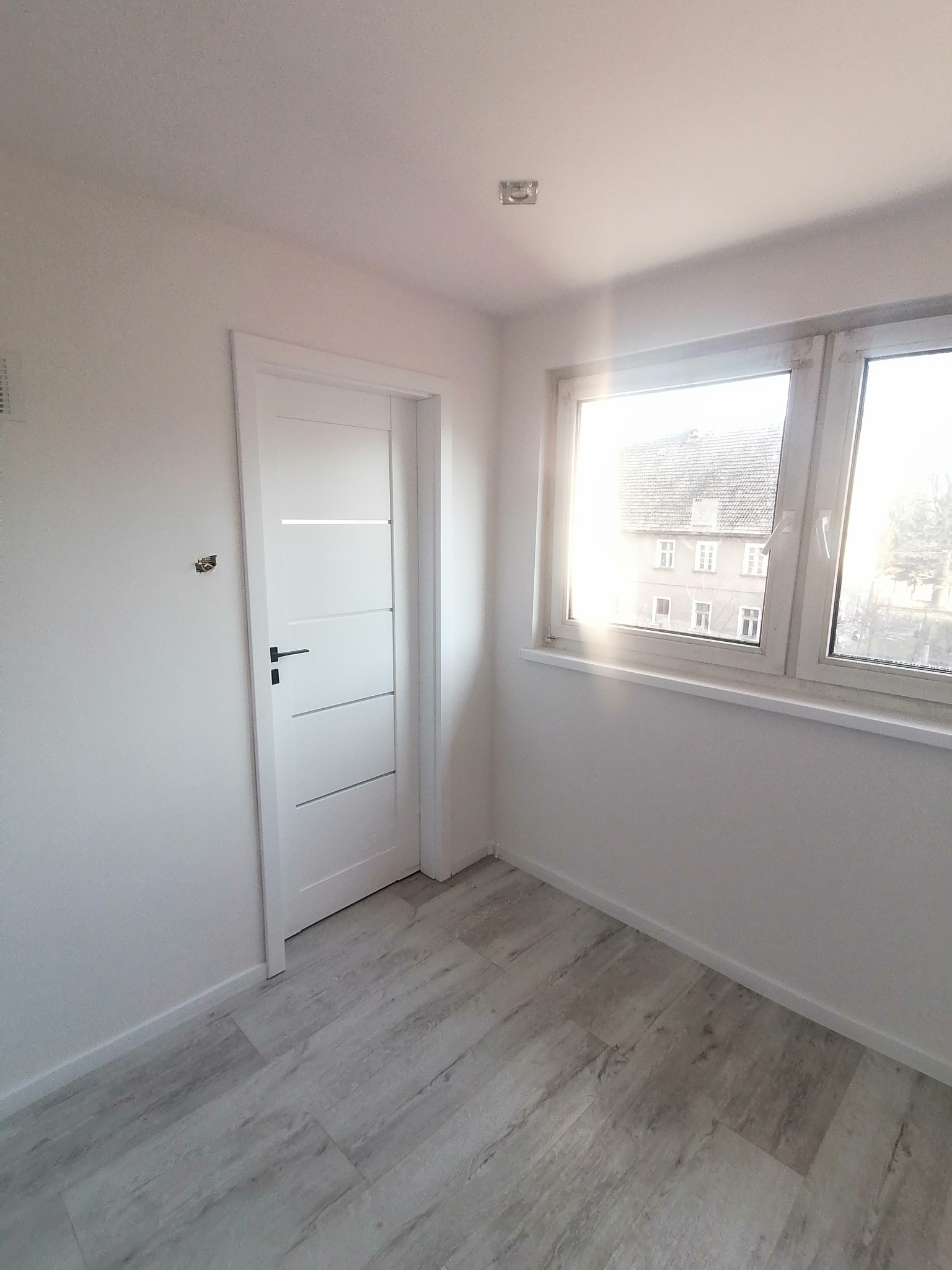 Wnętrze pokoju po remoncie: białe ściany, szare panele podłogowe imitujące drewno, białe drzwi z czarną klamką i okno z widokiem na budynek z czerwonej cegły.