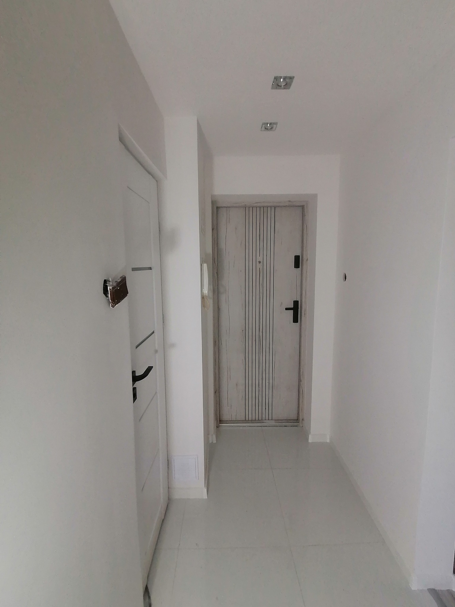 Biały korytarz z dwiema parami drzwi: wewnętrzne białe z czarną klamką i zewnętrzne w odcieniach szarości z pionowymi paskami, oświetlony kwadratowymi lampami sufitowymi.