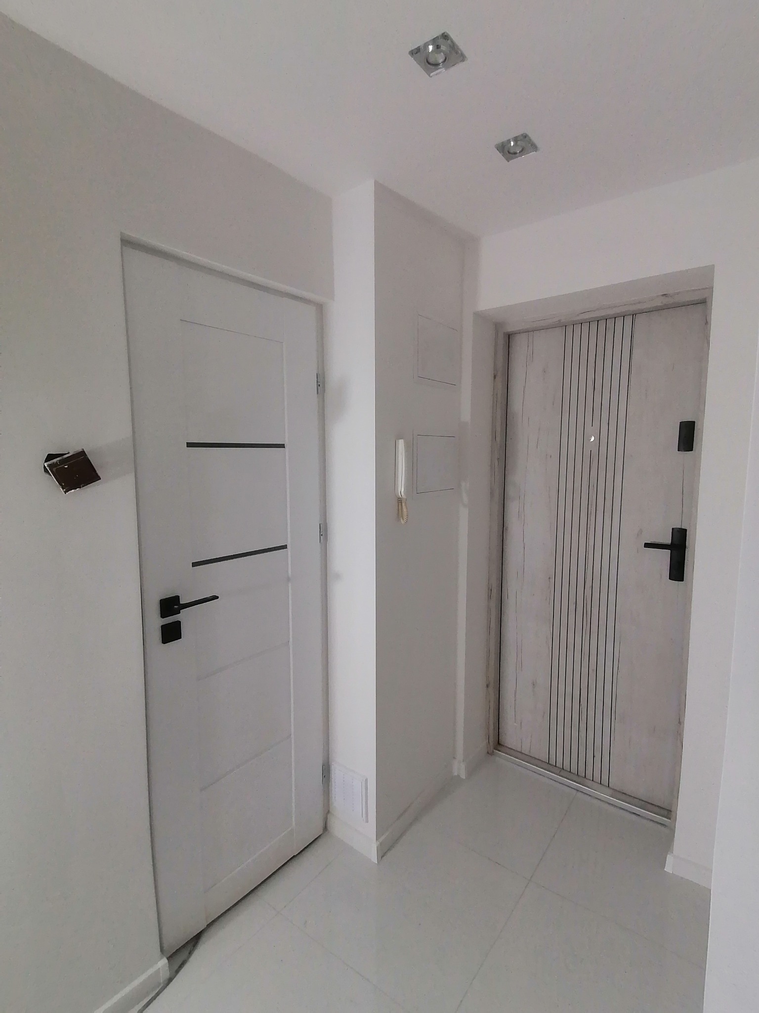 Nowoczesny, biały przedpokój z dwiema parami drzwi: wewnętrzne z poziomymi frezami i czarną klamką oraz zewnętrzne, drewnopodobne, z pionowymi żłobieniami i wizjerem.