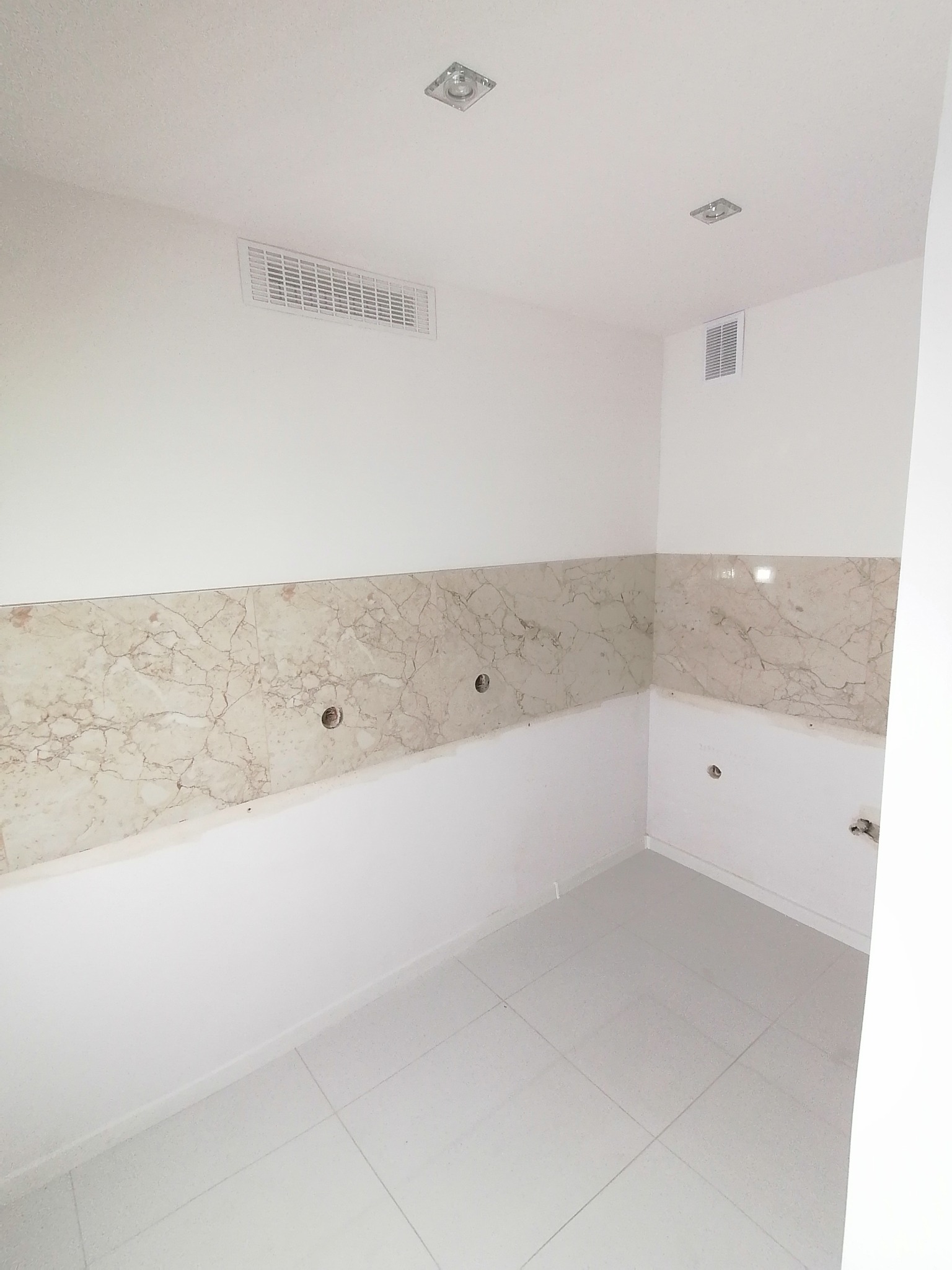 Wnętrze pomieszczenia w trakcie remontu, z położoną częściowo marmurową glazurą na ścianach, widoczne otwory na instalacje, białe ściany i podłoga z jasnych płytek, kratka wentylacyjna pod sufitem.