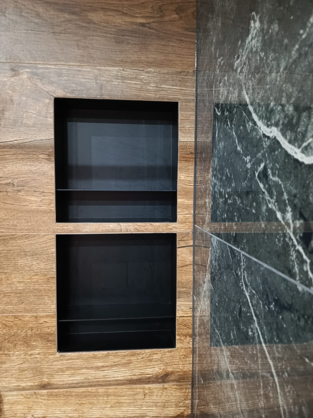 Wnęki w ścianie wyłożonej panelami drewnopodobnymi i płytami imitującymi marmur, z czarnym wnętrzem, tworzące nowoczesny element dekoracyjny.