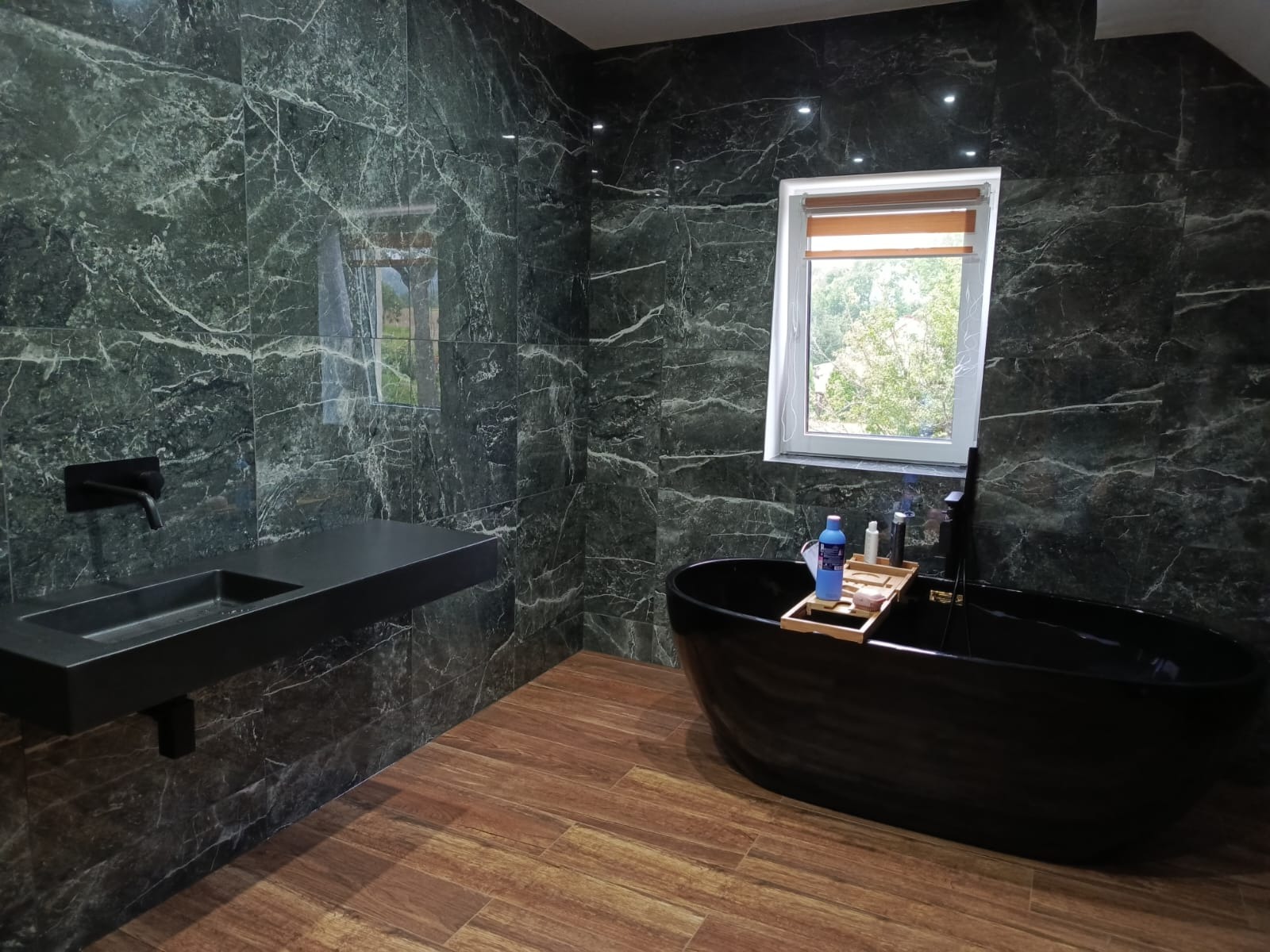 Elegancka łazienka z czarną wanną wolnostojącą i umywalką na tle ścian wyłożonych zielonym marmurem z białymi żyłami, drewnopodobna podłoga, okno z roletą w tle.