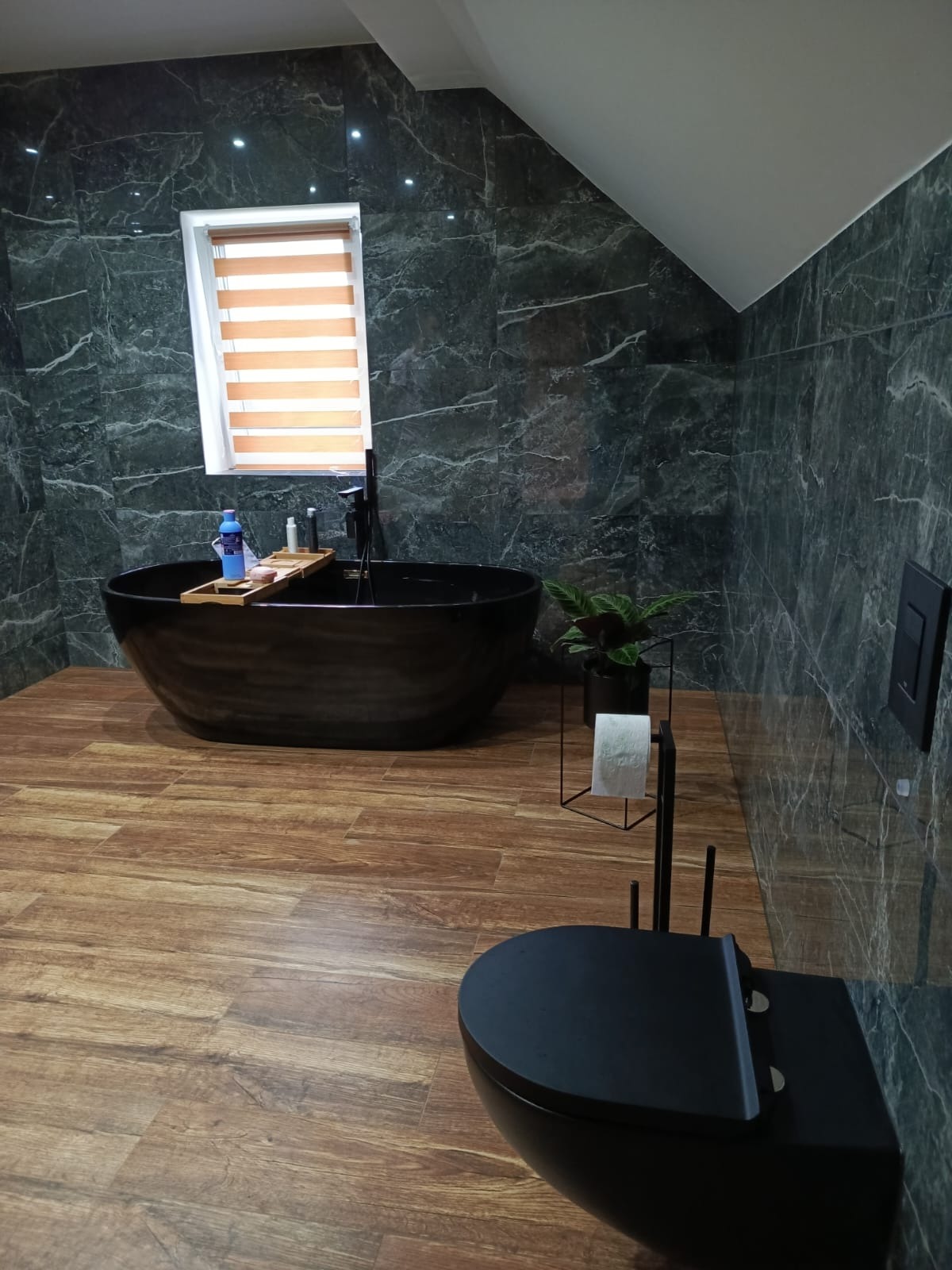 Nowoczesna łazienka na poddaszu z czarną wanną wolnostojącą, toaletą w tym samym kolorze, marmurowe ściany i drewnopodobna podłoga oraz rolety w oknie.