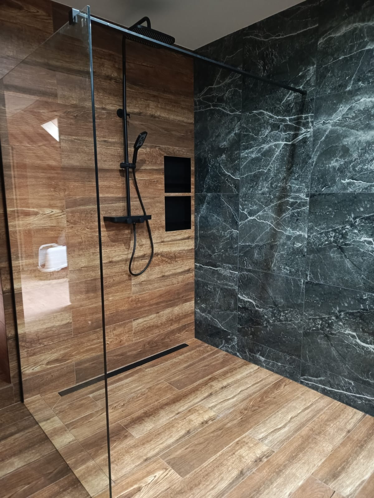 Nowoczesna kabina prysznicowa walk-in z czarną armaturą, wykończona płytkami imitującymi drewno i marmur, z odpływem liniowym w podłodze.