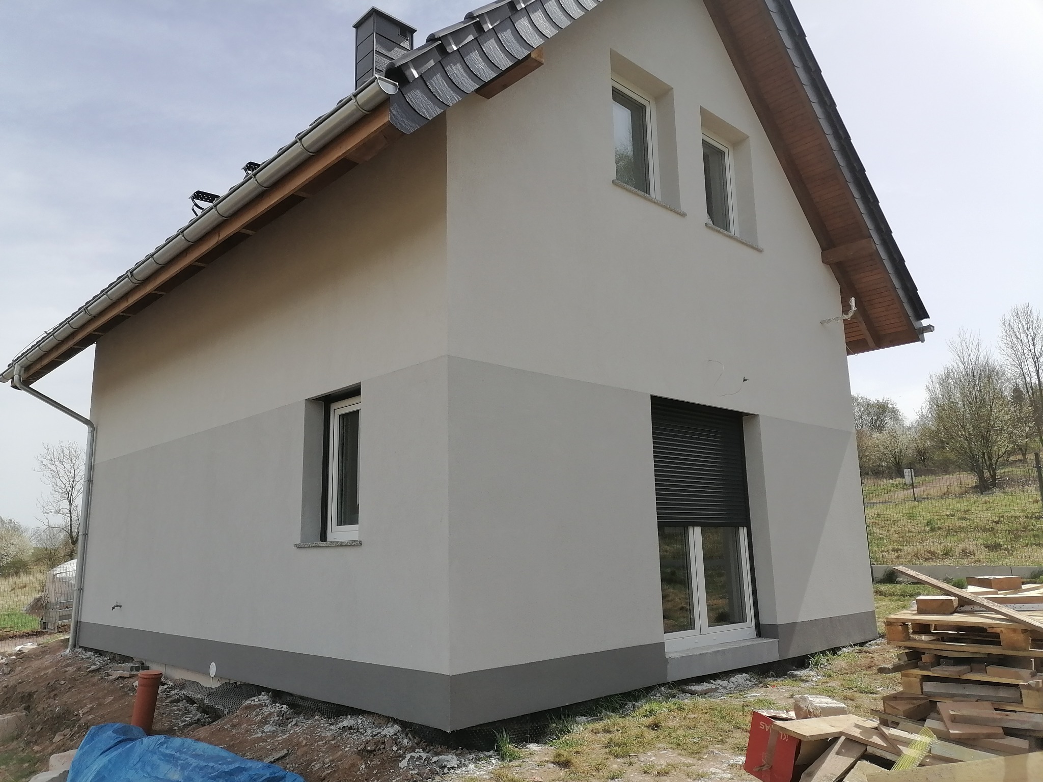 Nowoczesny dom jednorodzinny z jasną elewacją w dwóch odcieniach szarości, dach pokryty ciemną dachówką, okno z roletą zewnętrzną, widoczne materiały budowlane wokół domu.