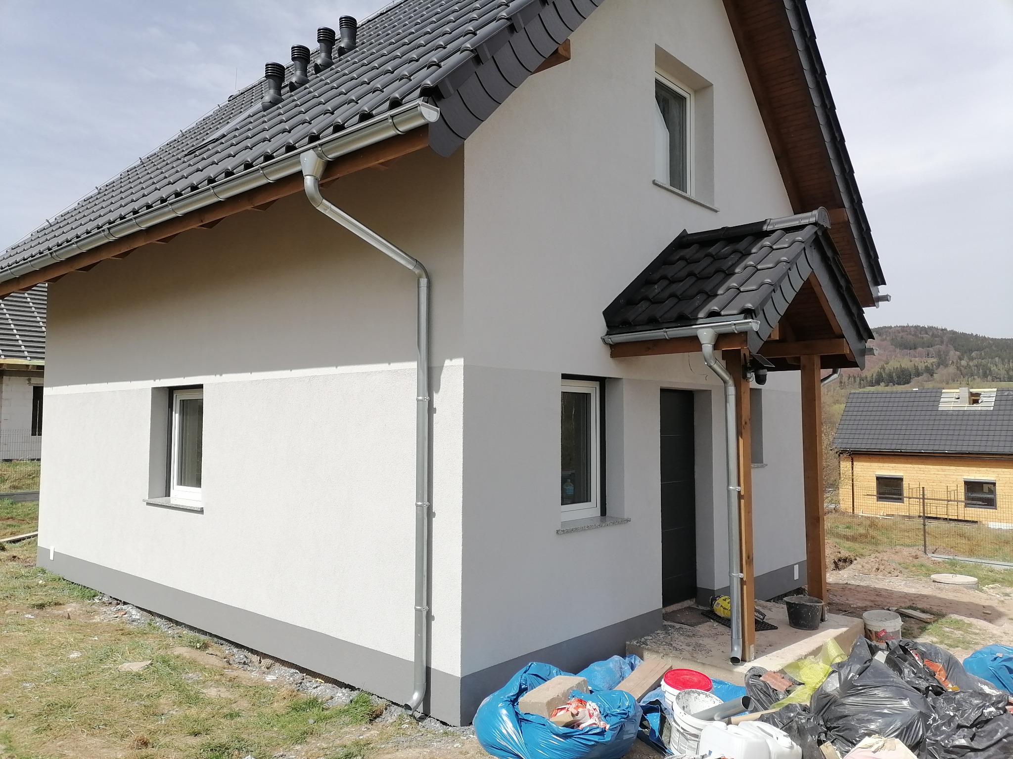 Nowo otynkowany dom jednorodzinny z szarą dachówką i orynnowaniem, widoczne worki z odpadami budowlanymi na trawniku.