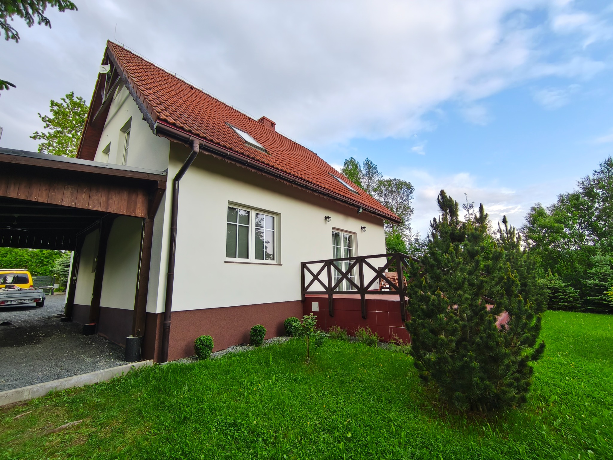 Biały dom z czerwonym dachem i drewnianą wiatą garażową, widok z zewnątrz, zieleń wokół. Widoczne okna, balustrada i elementy wykończeniowe.