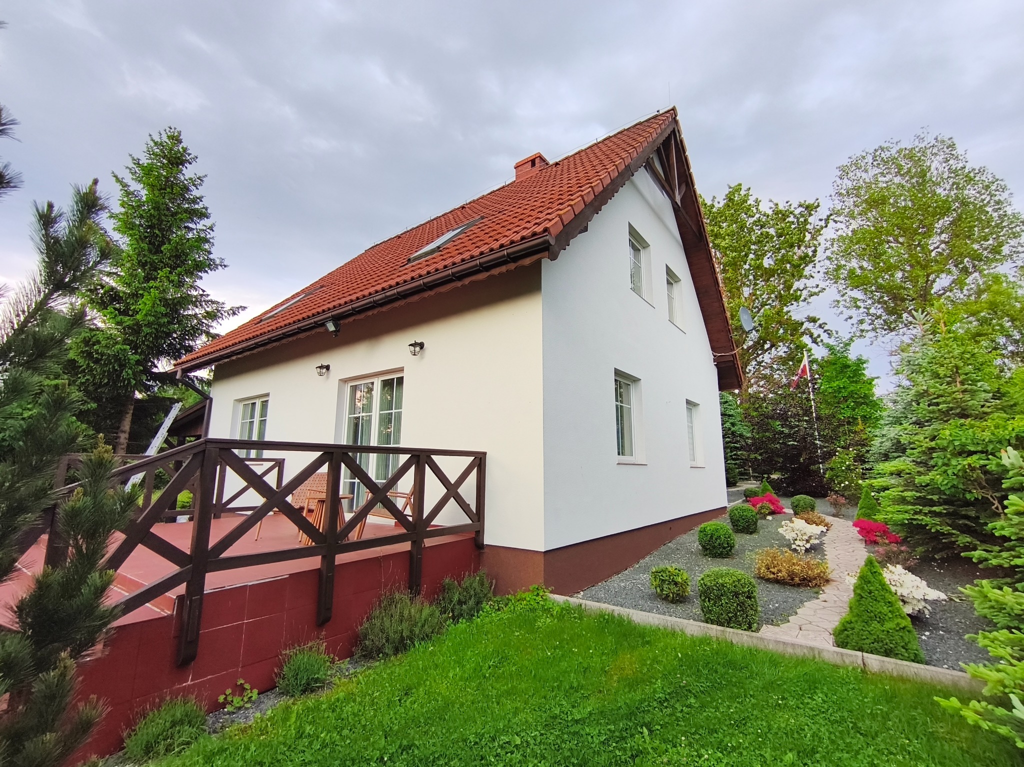 Biały dom z czerwoną dachówką i drewnianym tarasem, otoczony zielenią. Uwagę przykuwa świeżo odmalowana elewacja i zadbany ogród z ozdobnymi krzewami.