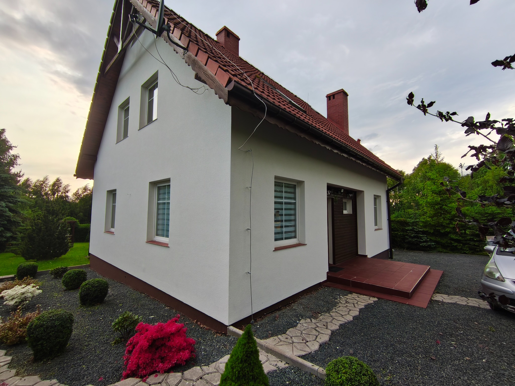 Biały dom z czerwoną dachówką i kominem, otoczony ozdobnym ogrodem z krzewami i ścieżką z kamieni. Widoczna elewacja po renowacji i nowy dach.