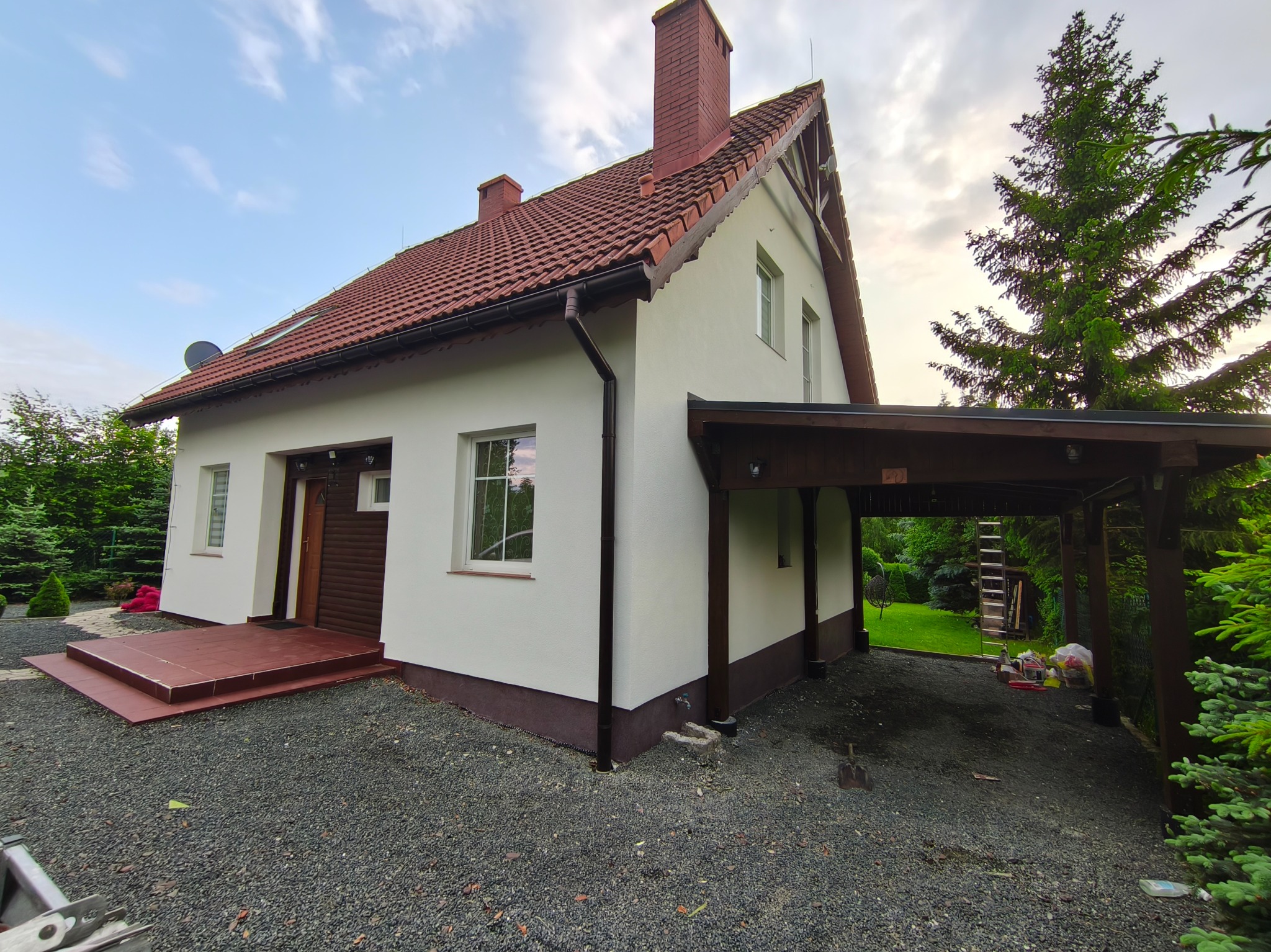 Jasny dom jednorodzinny z czerwoną dachówką i podjazdem z szarego żwiru. Widoczny fragment elewacji, okna, drzwi, zadaszenie na samochód i fragment ogrodu.