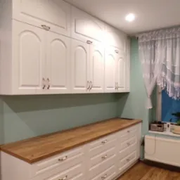 Białe szafki kuchenne z frezowanymi frontami i drewnianym blatem, uzupełnione o białą przesuwną szafę wnękową, okno z białą firanką i roślinami doniczkowymi.