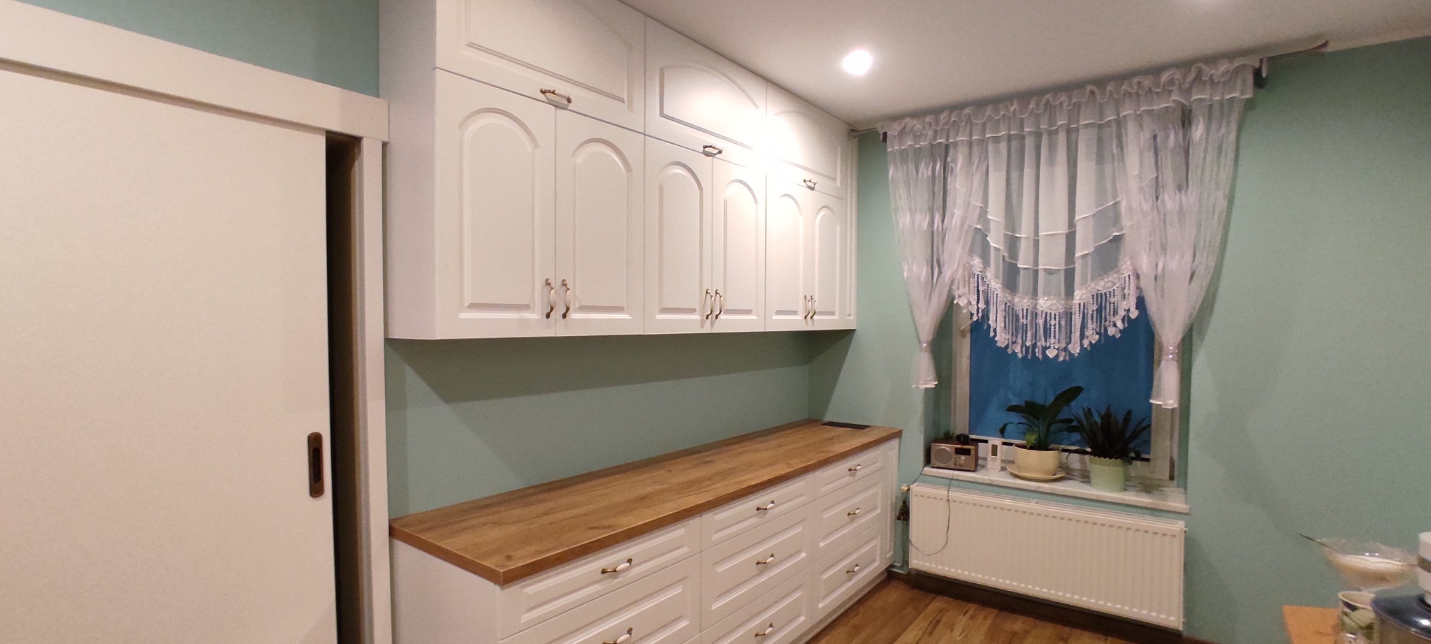 Białe szafki kuchenne z frezowanymi frontami i drewnianym blatem, uzupełnione o białą przesuwną szafę wnękową, okno z białą firanką i roślinami doniczkowymi.