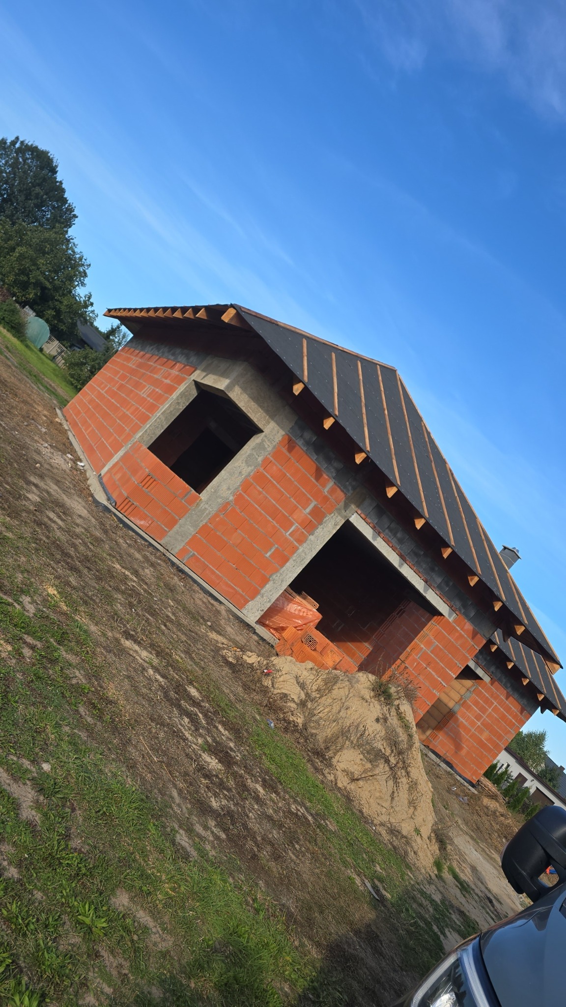 Niedokończony budynek z czerwonej cegły z widocznym dachem i otworami okiennymi, na tle błękitnego nieba. Widoczne materiały budowlane.