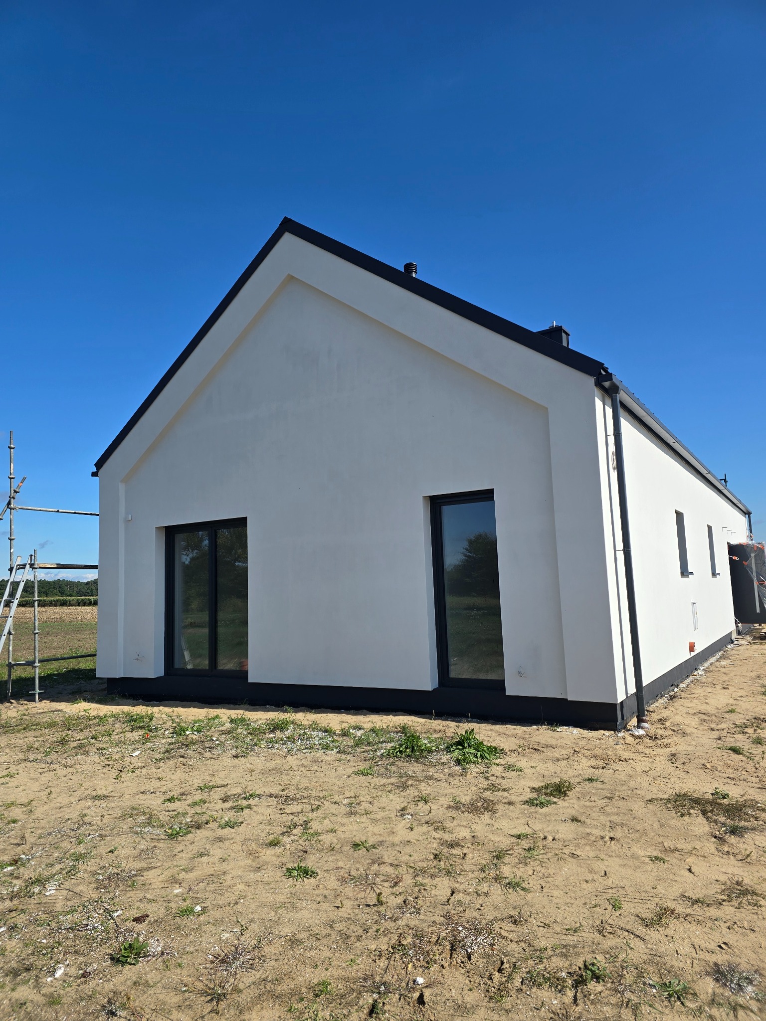 Nowoczesny, biały dom z czarnymi ramami okiennymi i dachem na tle błękitnego nieba. Widoczne rusztowania z lewej strony sugerują prace wykończeniowe.