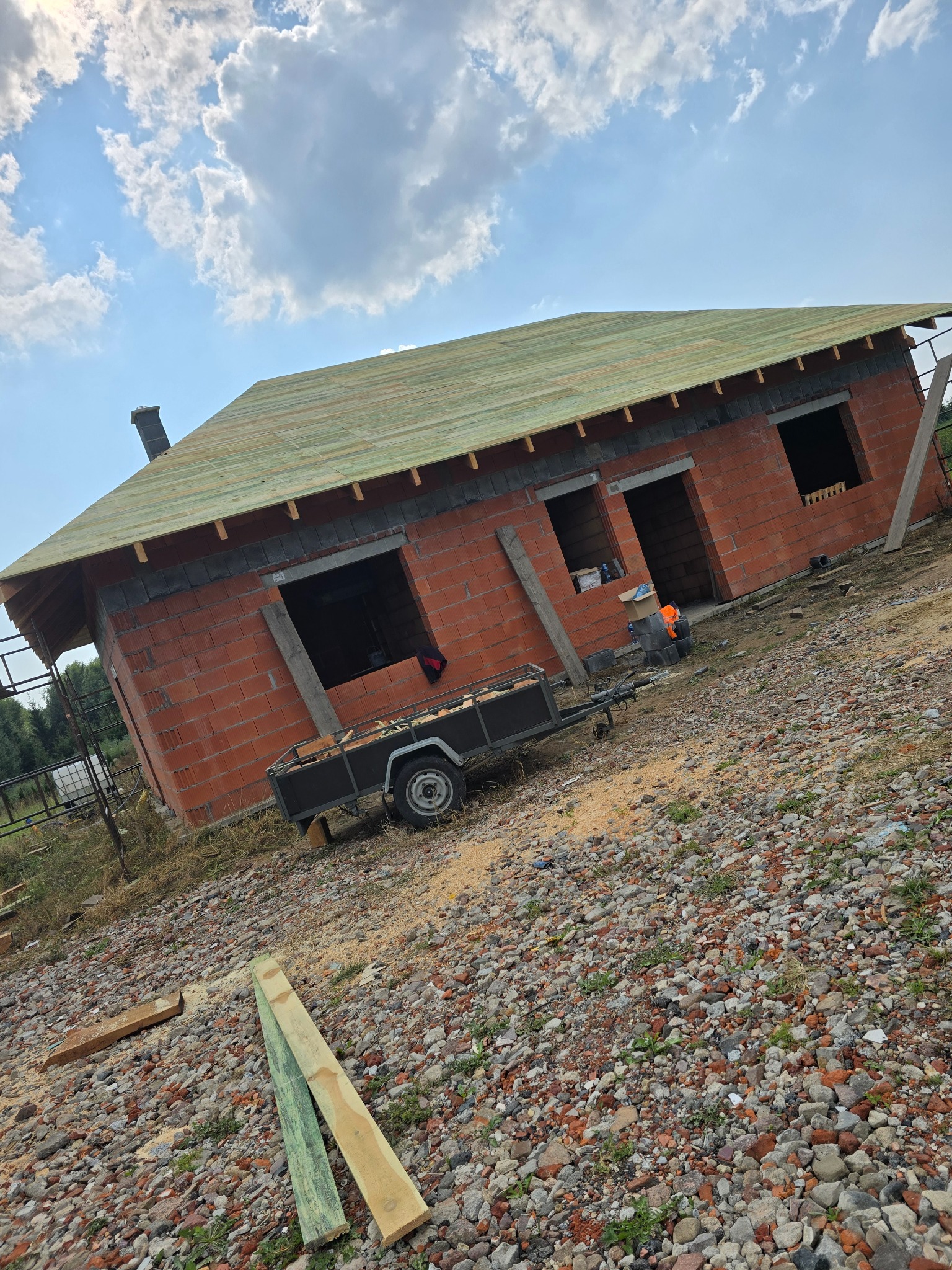 Niedokończony dom z czerwonej cegły z drewnianym dachem, przyczepą i gruzem na działce w słoneczny dzień. Widoczne otwory okienne i komin.