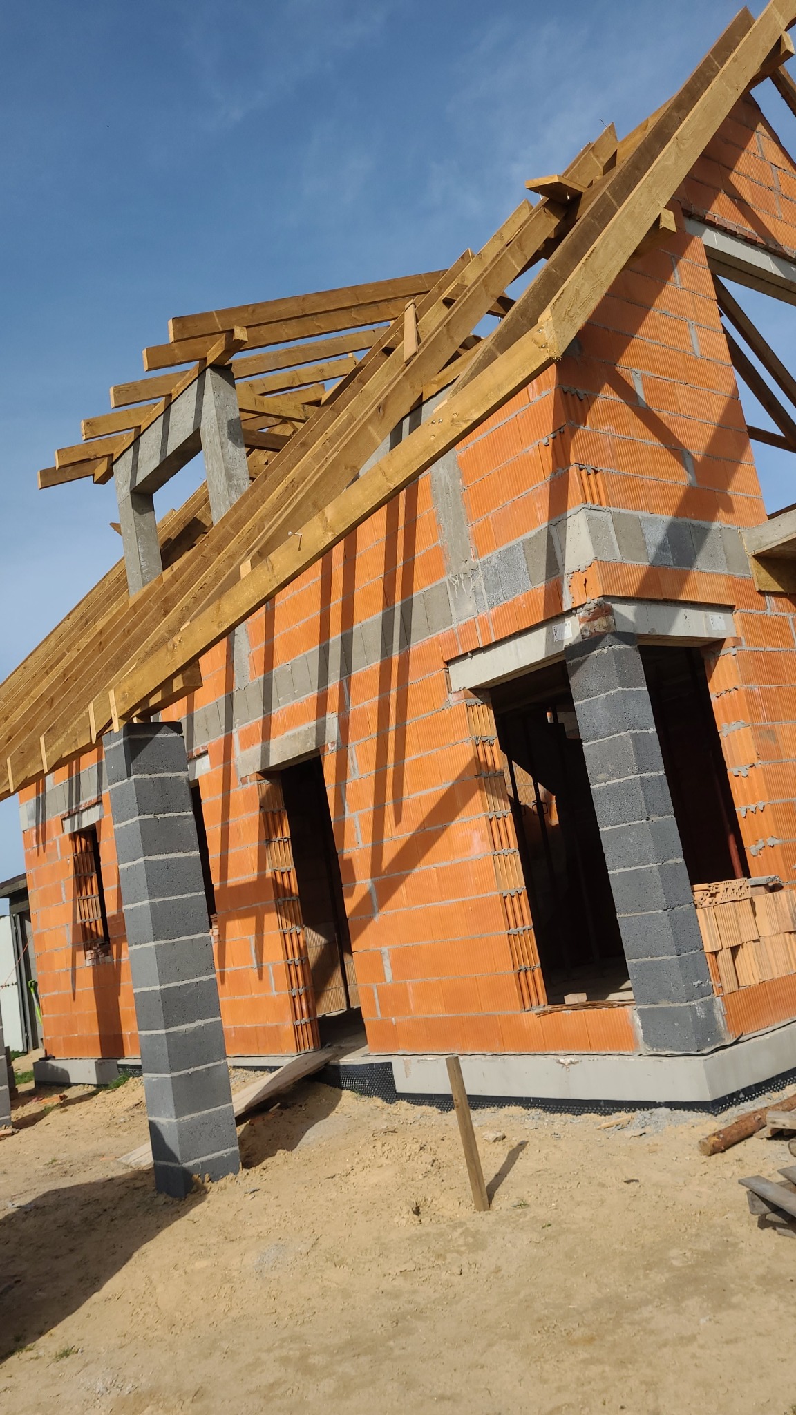 Surowy stan domu z czerwonej cegły z widoczną konstrukcją dachu z drewna, na tle błękitnego nieba. Widoczne otwory okienne i drzwiowe.