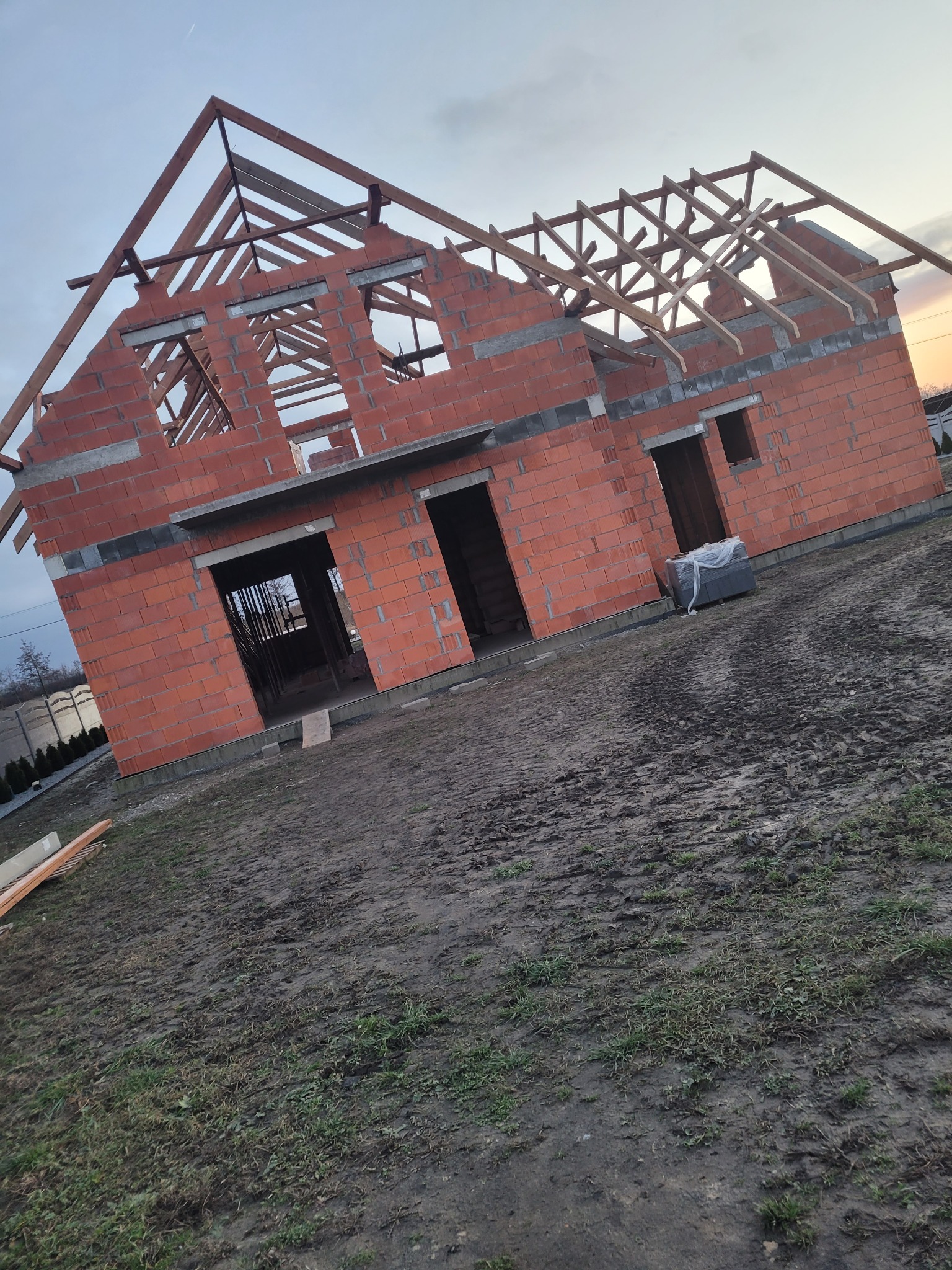 Surowy stan domu z czerwonej cegły z drewnianą konstrukcją dachu w trakcie budowy, na tle pochmurnego nieba o zmierzchu. Widoczne ślady opon na błotnistym terenie.