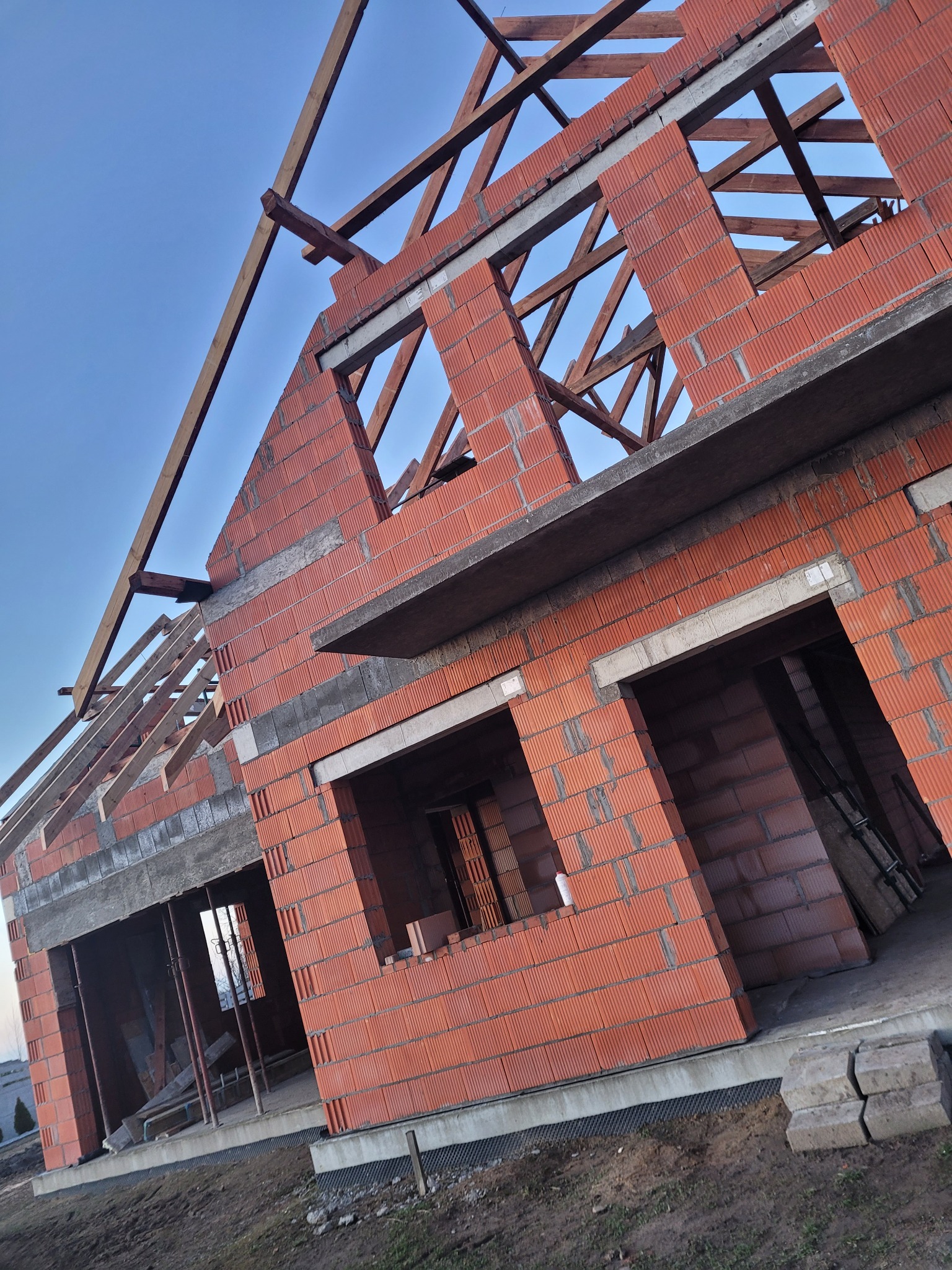 Surowy stan domu z czerwonej cegły, widok pod kątem, z widoczną konstrukcją dachu i otworami okiennymi, w tle błękitne niebo. Etap budowy domu.