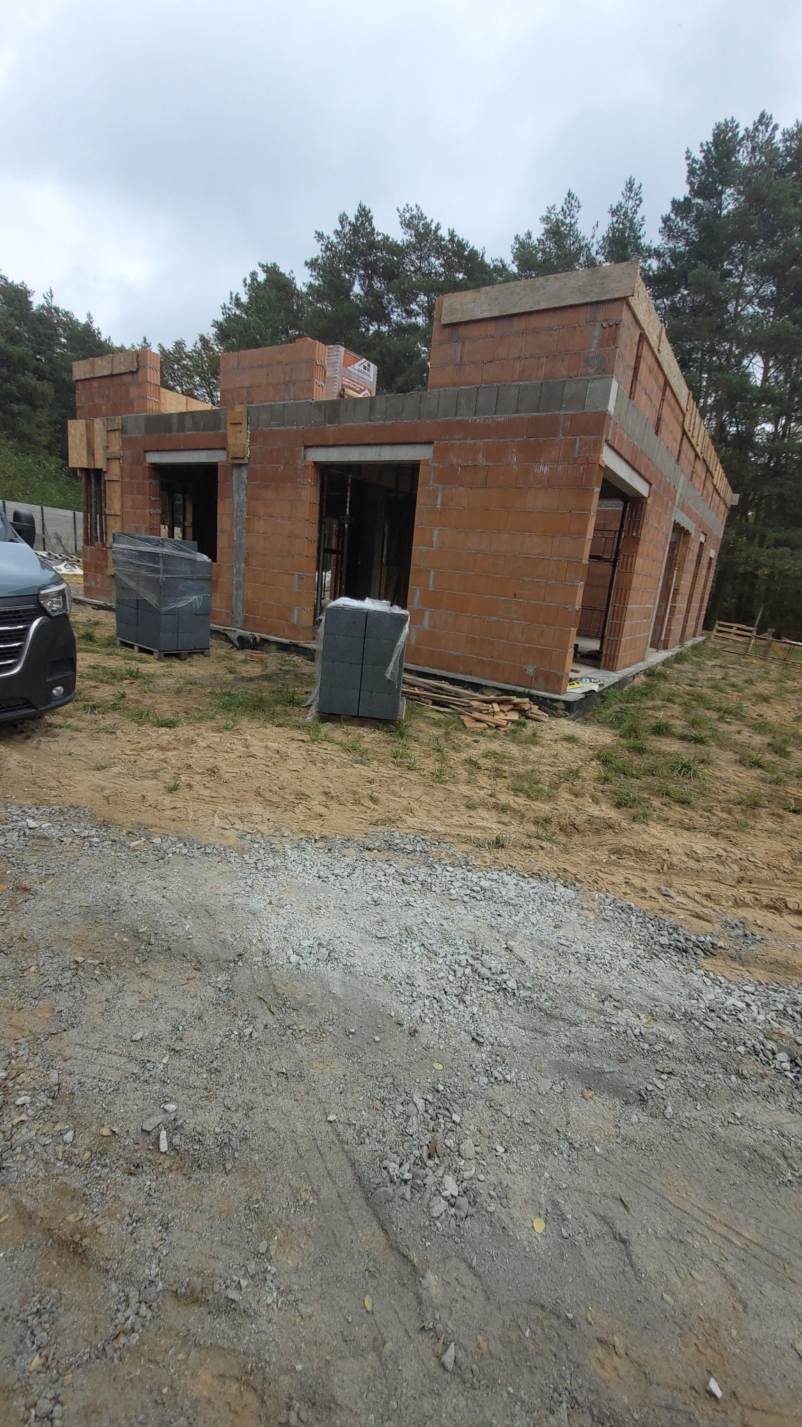 Budowa domu w trakcie: ściany z czerwonej cegły, widoczne otwory okienne i drzwiowe, palety z materiałami budowlanymi obok. W tle las i pochmurne niebo.