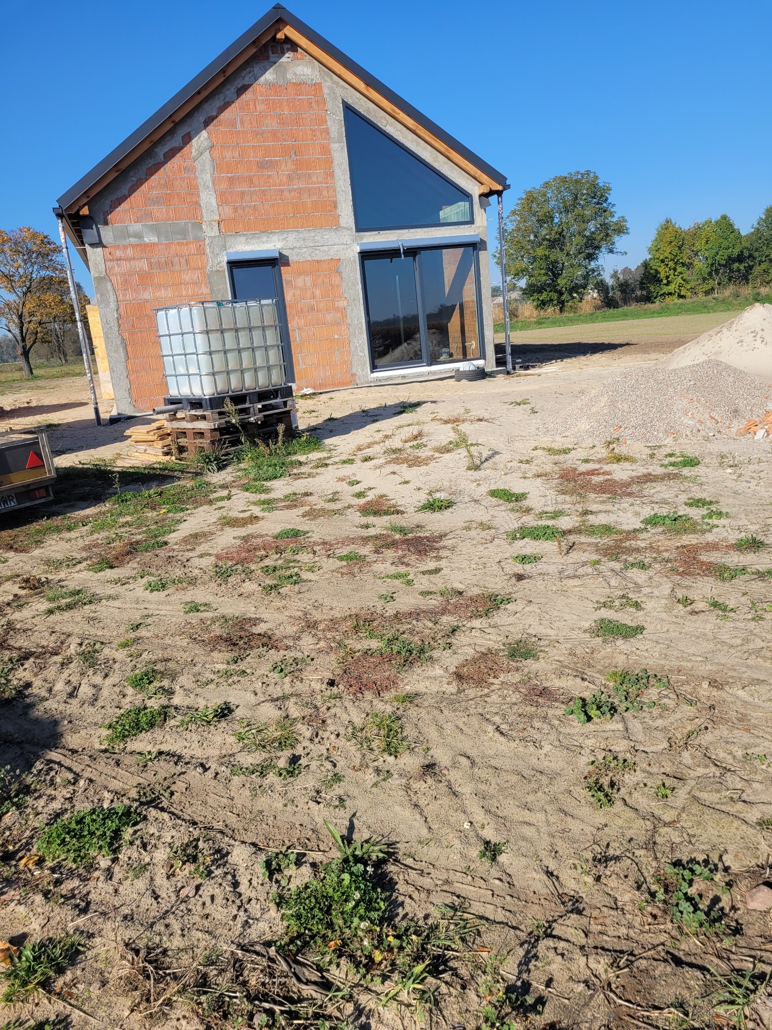 Surowy stan domu z czerwonej cegły z dużym oknem, zbiornik na wodę na paletach obok, teren budowy z piaskiem i zielenią, błękitne niebo w tle.
