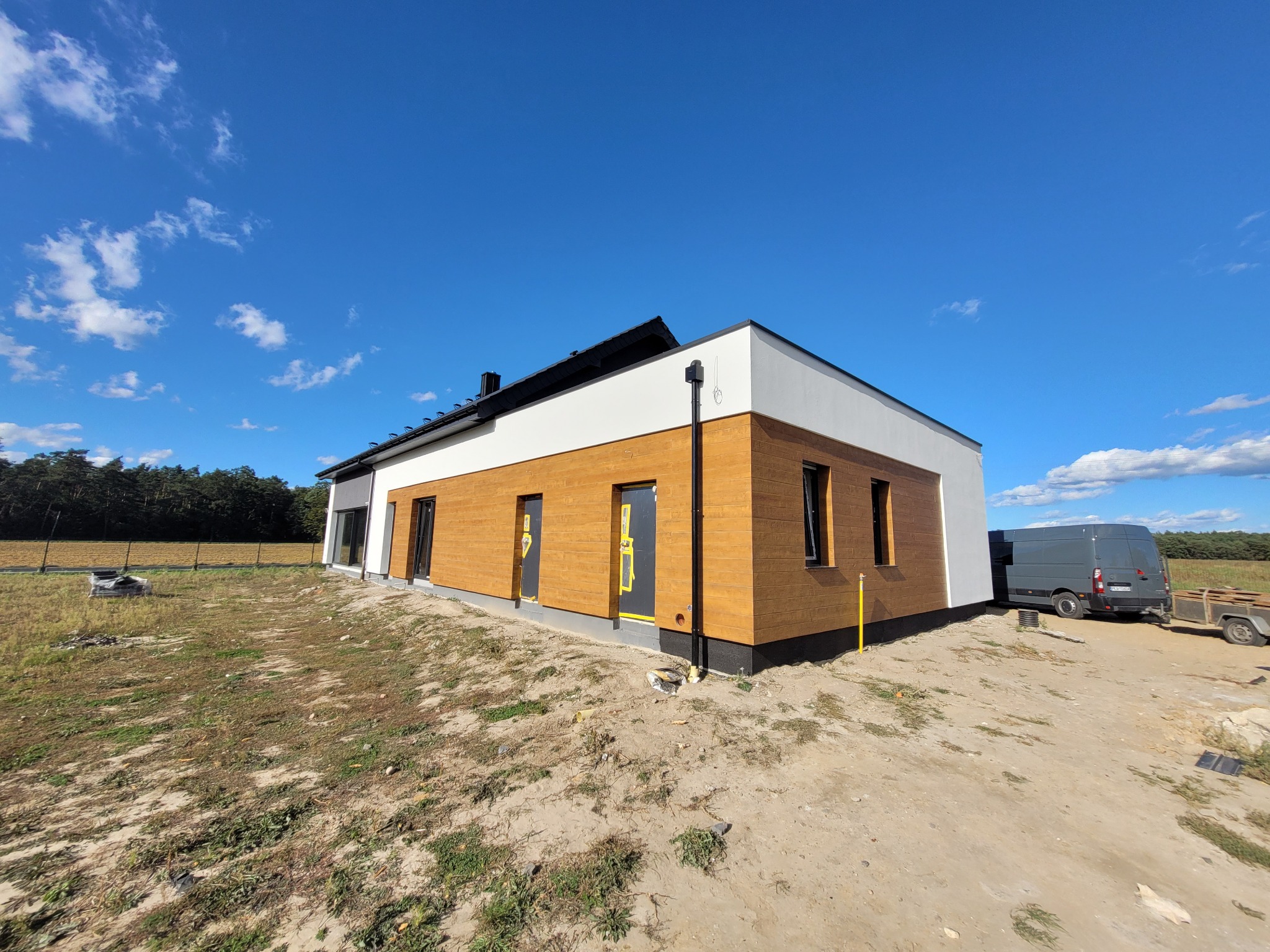 Nowoczesny dom jednorodzinny z drewnianą elewacją i białym wykończeniem, na tle błękitnego nieba z chmurami. Widoczny fragment działki i samochód dostawczy w tle.