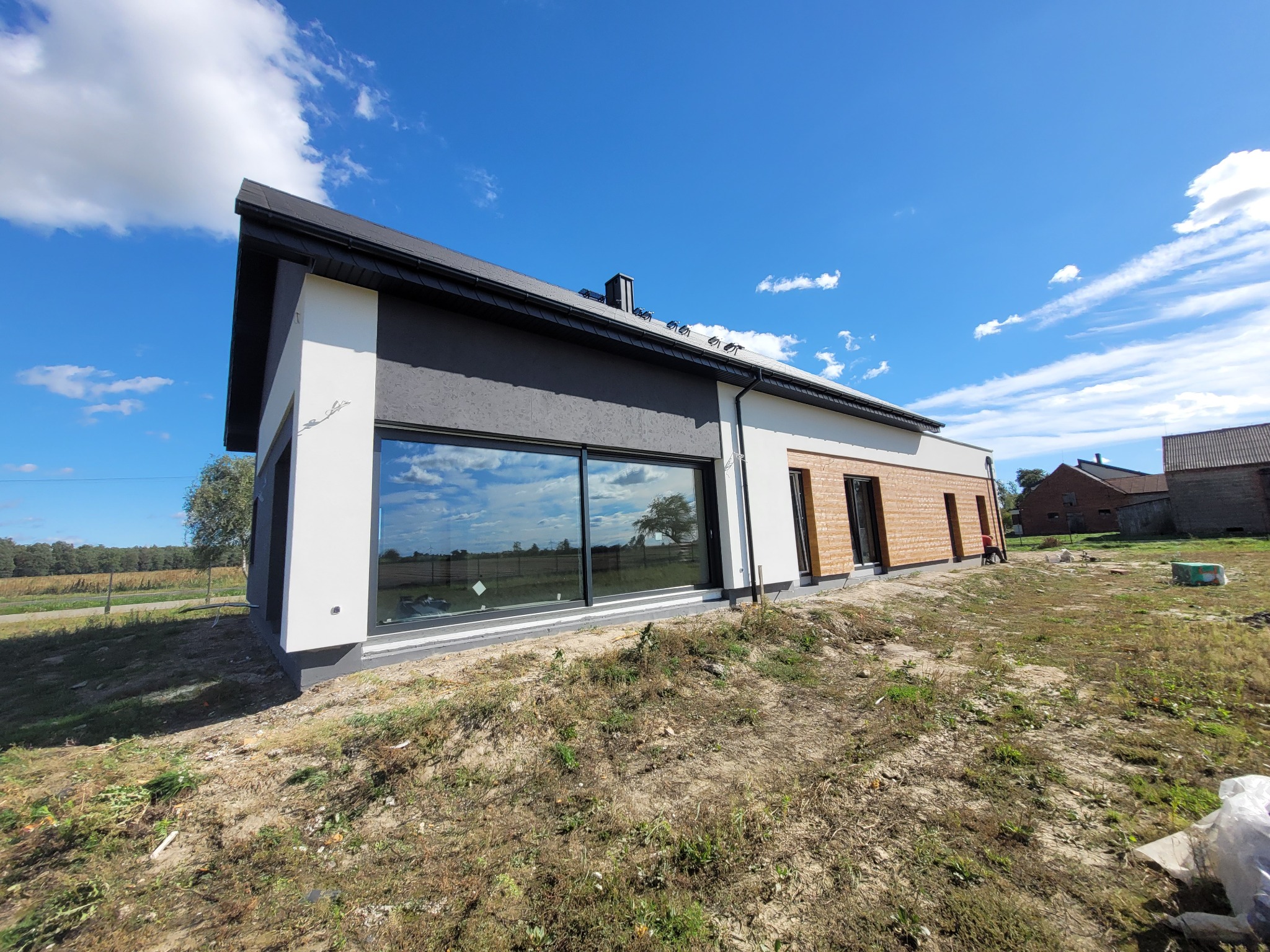 Nowoczesny dom jednorodzinny z dużym oknem, wykończony w bieli i szarości, z elementami drewna. Widok z zewnątrz, w tle błękitne niebo z chmurami i zieleń drzew.