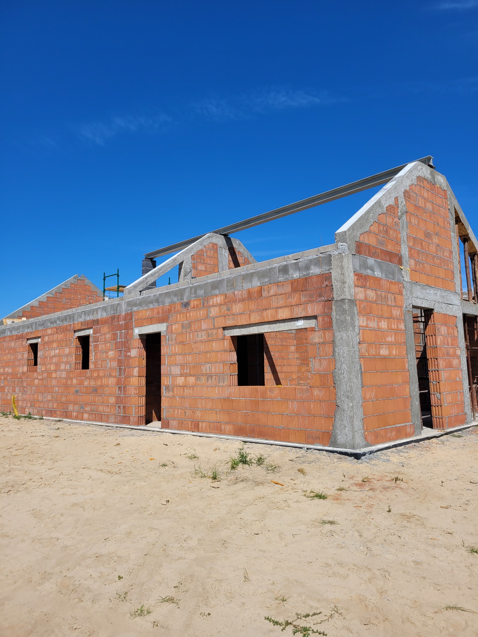 Budowa domu z czerwonej cegły, widoczne otwory okienne i drzwiowe, stalowe belki dachowe, błękitne niebo w tle. Stan surowy otwarty.