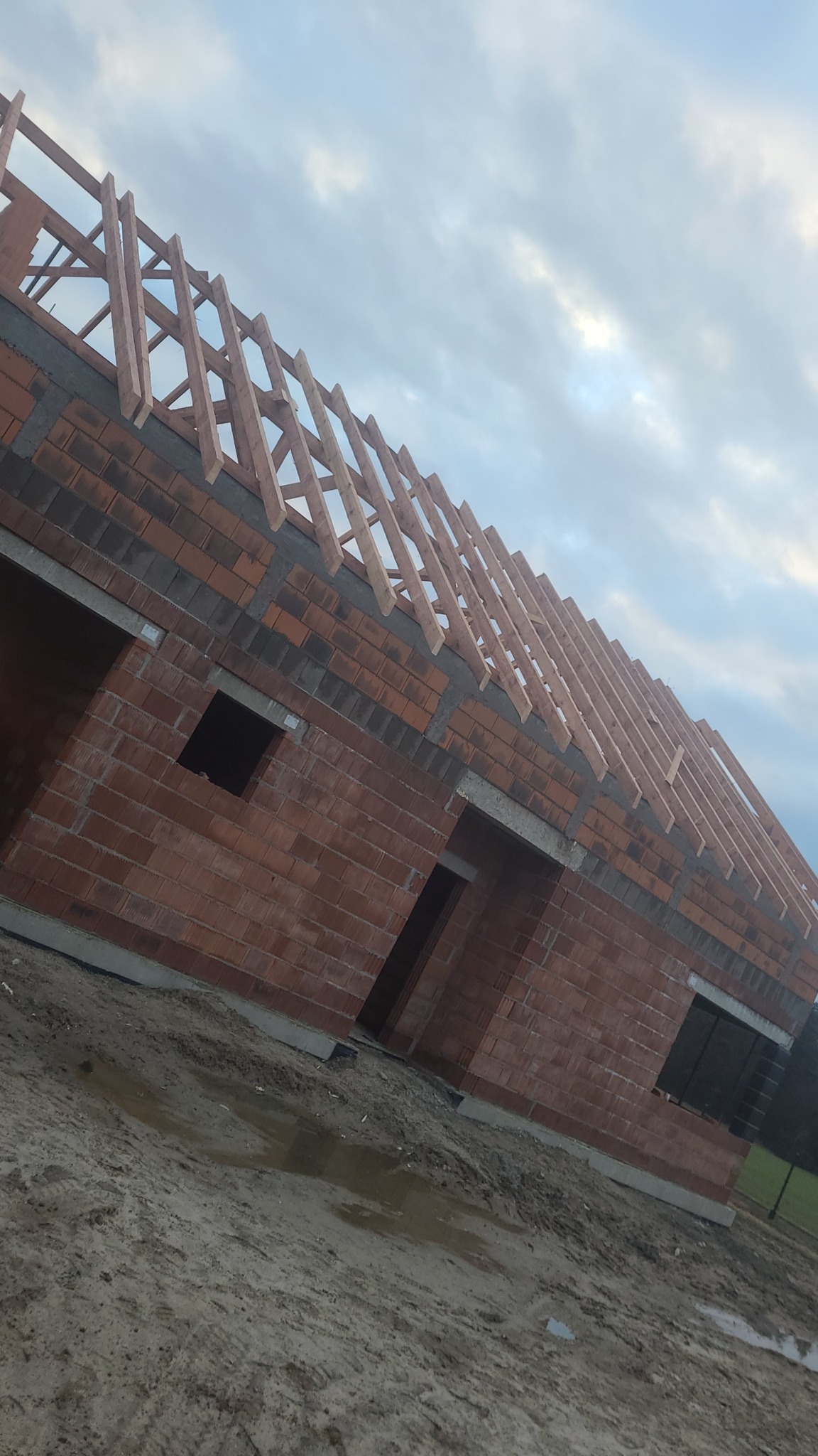 Surowy stan domu z czerwonej cegły z drewnianą konstrukcją dachu w trakcie budowy, widok z dołu na tle pochmurnego nieba.