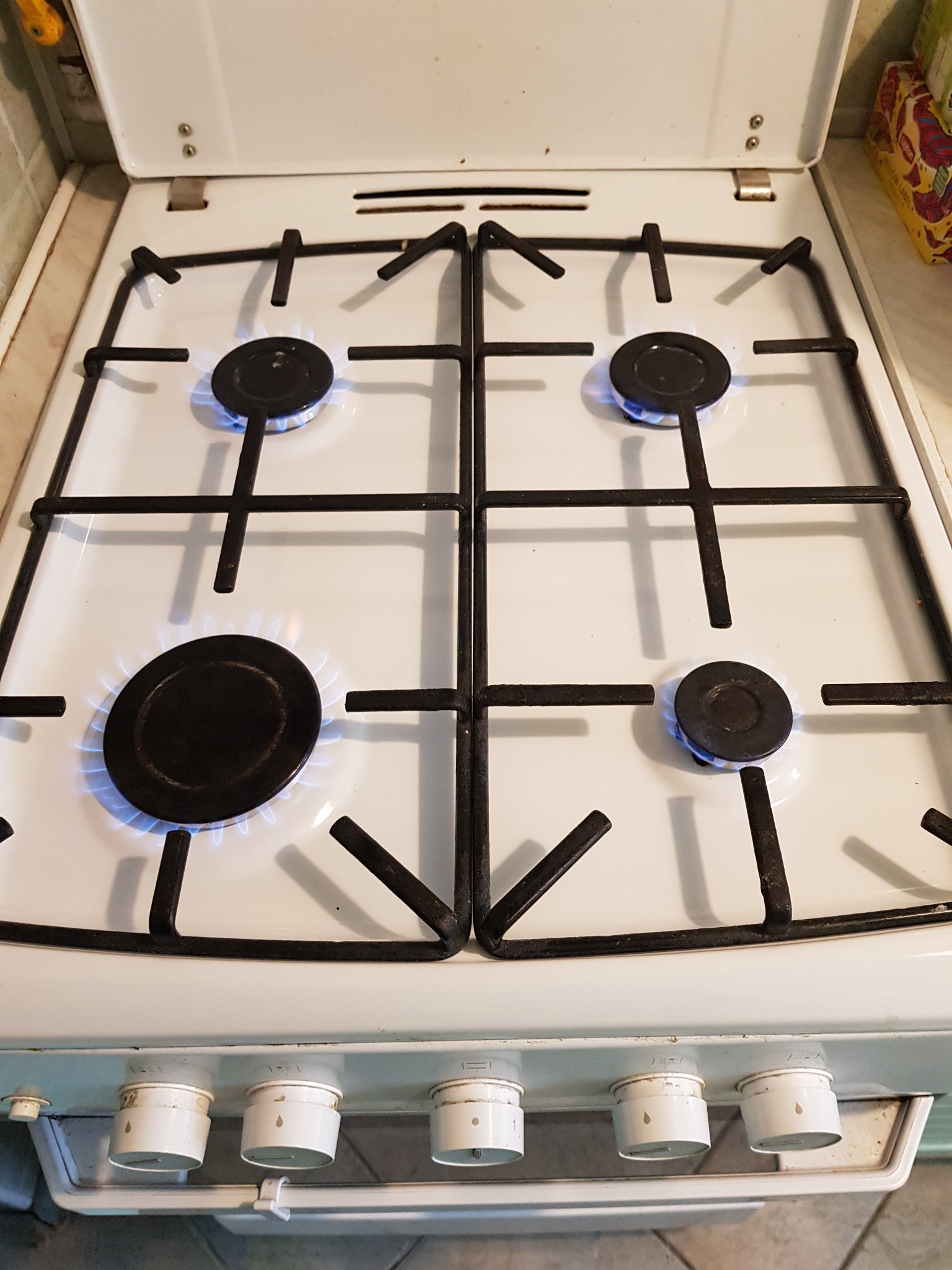 Biała, używana kuchenka gazowa z otwartą klapą, włączone cztery palniki z widocznym niebieskim płomieniem, emaliowane ruszty nad palnikami, widoczne pokrętła regulacji gazu.