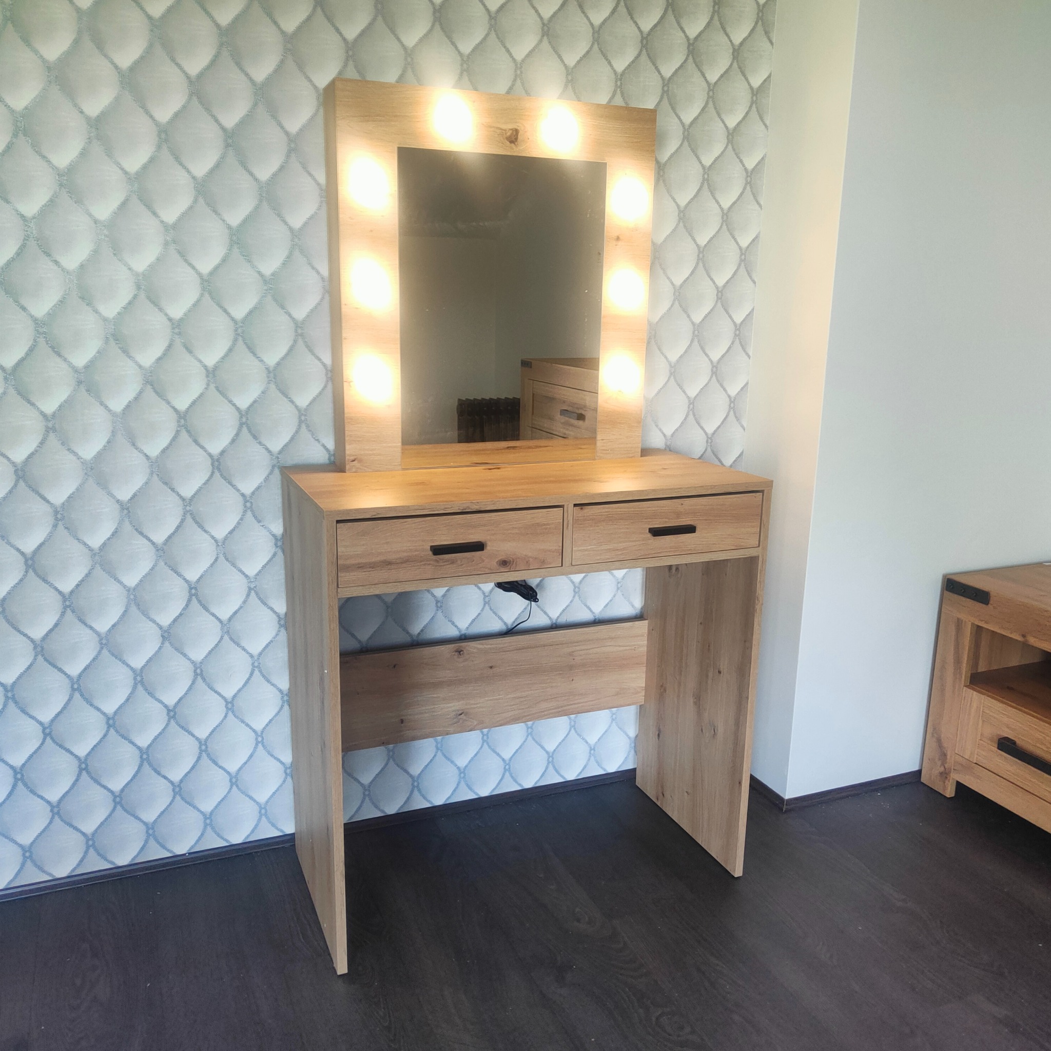 Drewniana toaletka z podświetlanym lustrem i dwoma szufladami, ustawiona na ciemnej podłodze na tle wzorzystej tapety.