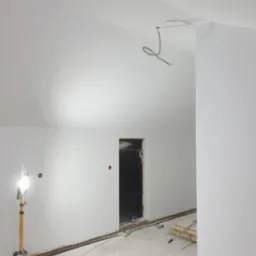 Poddasze w trakcie remontu: białe ściany po gładzi, otwór drzwiowy bez ościeżnicy, wystające przewody elektryczne i halogen na statywie oświetlający pomieszczenie, podłoga w trakcie prac.
