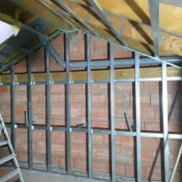 Poddasze w trakcie adaptacji: widoczna konstrukcja metalowa na tle ceglanej ściany, szara rura wentylacyjna biegnąca poziomo, drewniany strop i drabina przystawna z lewej strony.