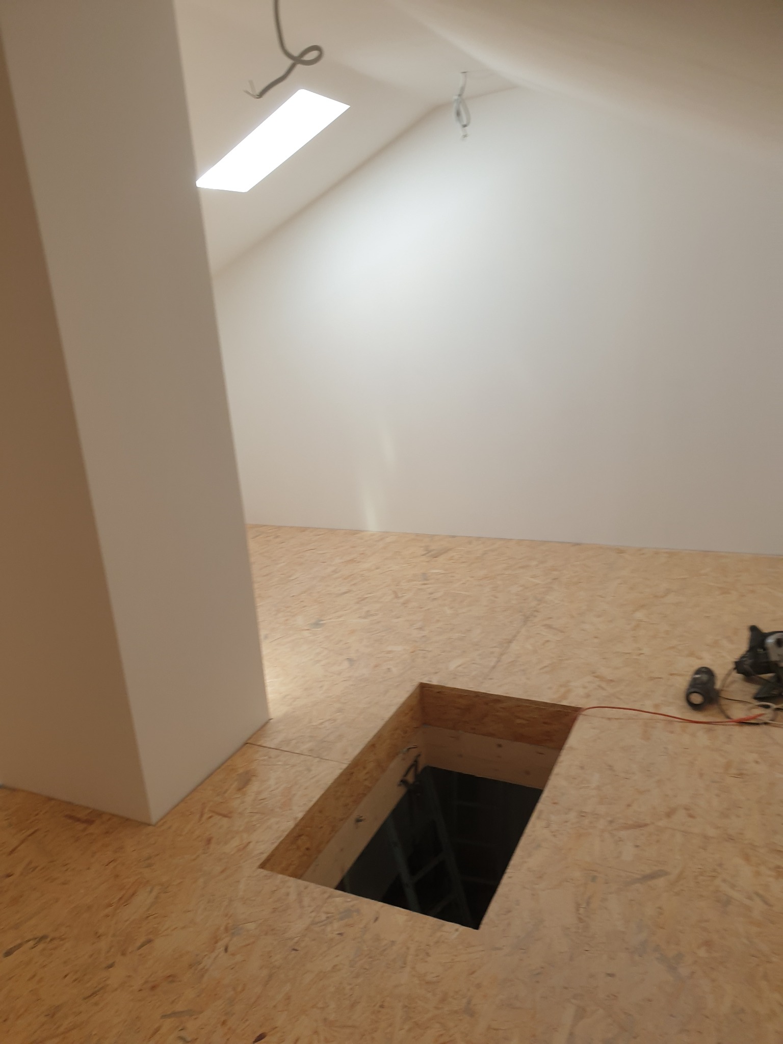 Poddasze z wyciętym otworem w podłodze z płyty OSB, widoczna drabina w otworze, narzędzia leżące obok, białe ściany i kolumna.