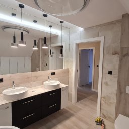 Grzegorz Szymoniak - Nowoczesna łazienka z dwoma okrągłymi umywalkami nablatowymi, czarnymi szafkami pod umywalkami i dużym lustrem z wiszącymi lampami, wykończona płytkami imitującymi drewno.