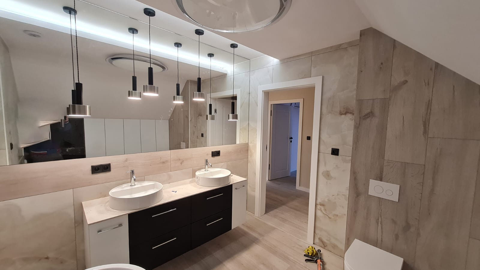 Nowoczesna łazienka z dwoma okrągłymi umywalkami nablatowymi, czarnymi szafkami pod umywalkami i dużym lustrem z wiszącymi lampami, wykończona płytkami imitującymi drewno.