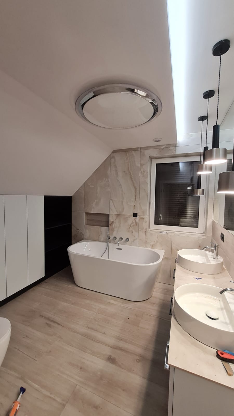 Nowoczesna łazienka na poddaszu z wolnostojącą wanną, dwiema okrągłymi umywalkami nablatowymi, marmurowymi płytkami na ścianach i drewnopodobną podłogą; widoczna zabudowa meblowa z białymi frontami...
