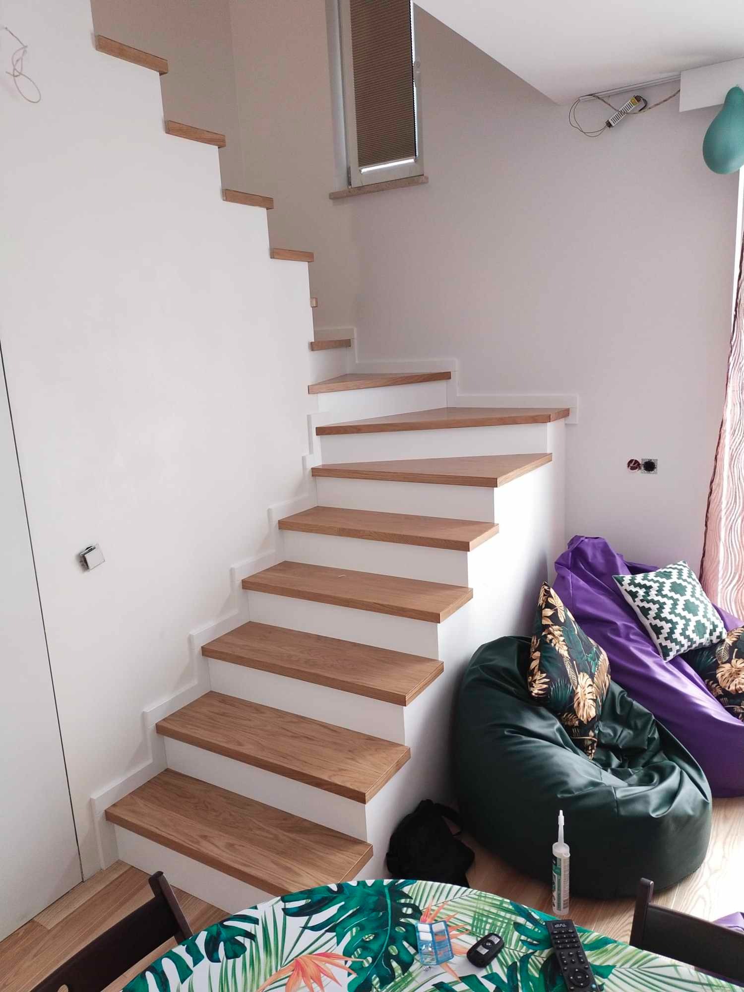 Białe schody z drewnianymi stopniami, prowadzące do góry, widoczne z dołu, z elementami dekoracyjnymi w tle, takimi jak pufy i stolik z tropikalnym wzorem.